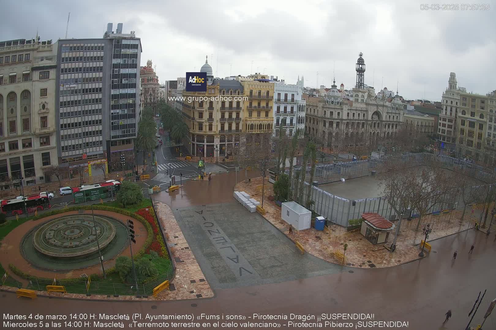 A wet city square with a fountain and several buildings is seen from an elevated view on an overcast day.