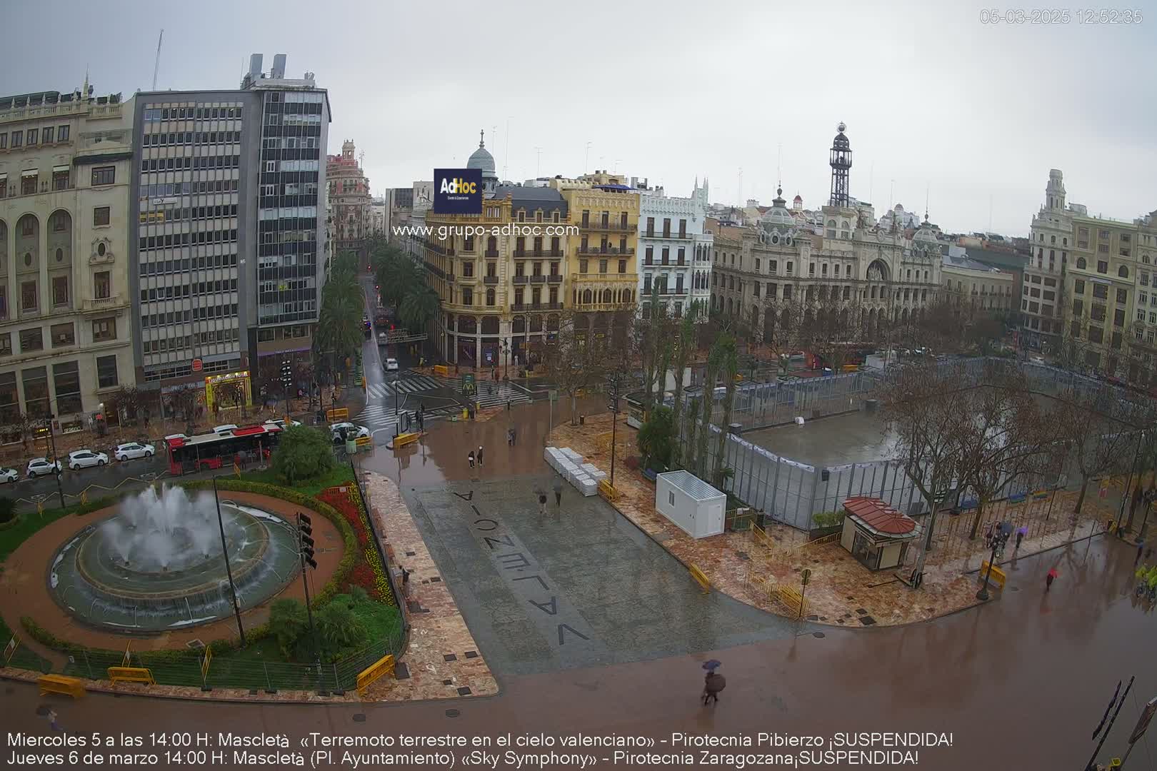 A city square, flooded by rain, features a large fountain, several buildings, and people walking under an overcast sky.