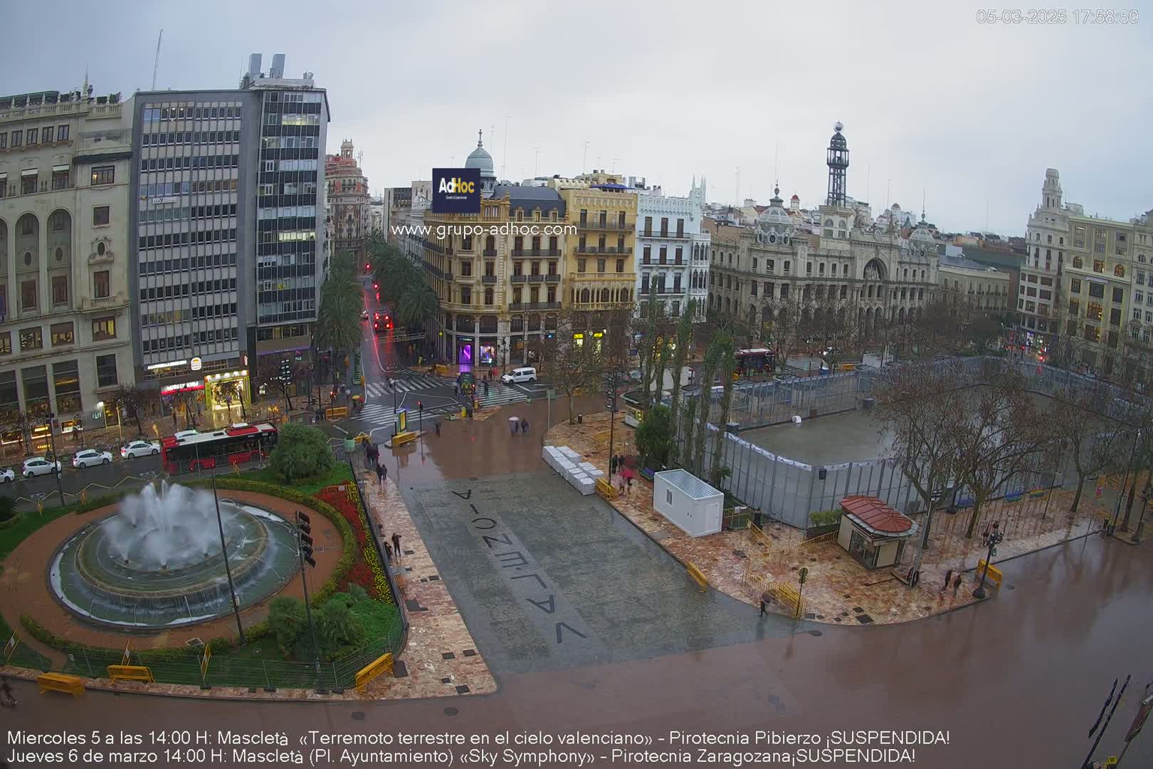 A city square, wet from rain, features a large fountain, buildings surrounding it, and a fenced-in area in the center.