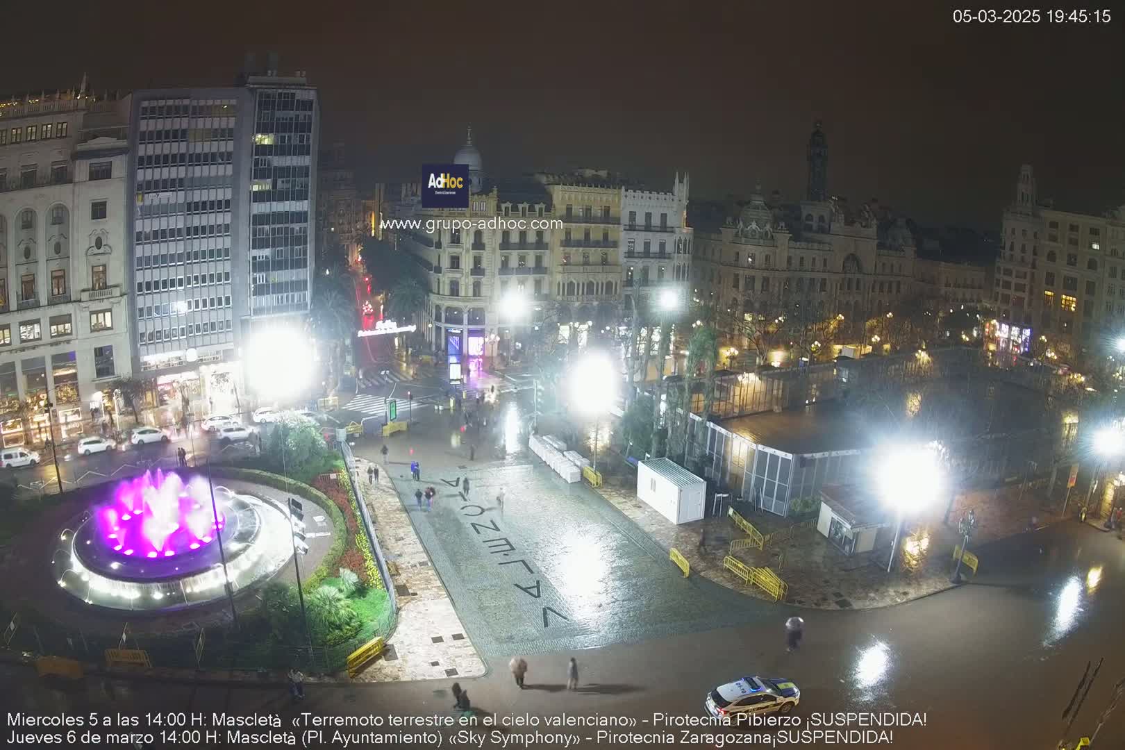 A nighttime, rain-soaked city square features a fountain with purple lights, buildings, and vehicles, all under bright artificial lighting.