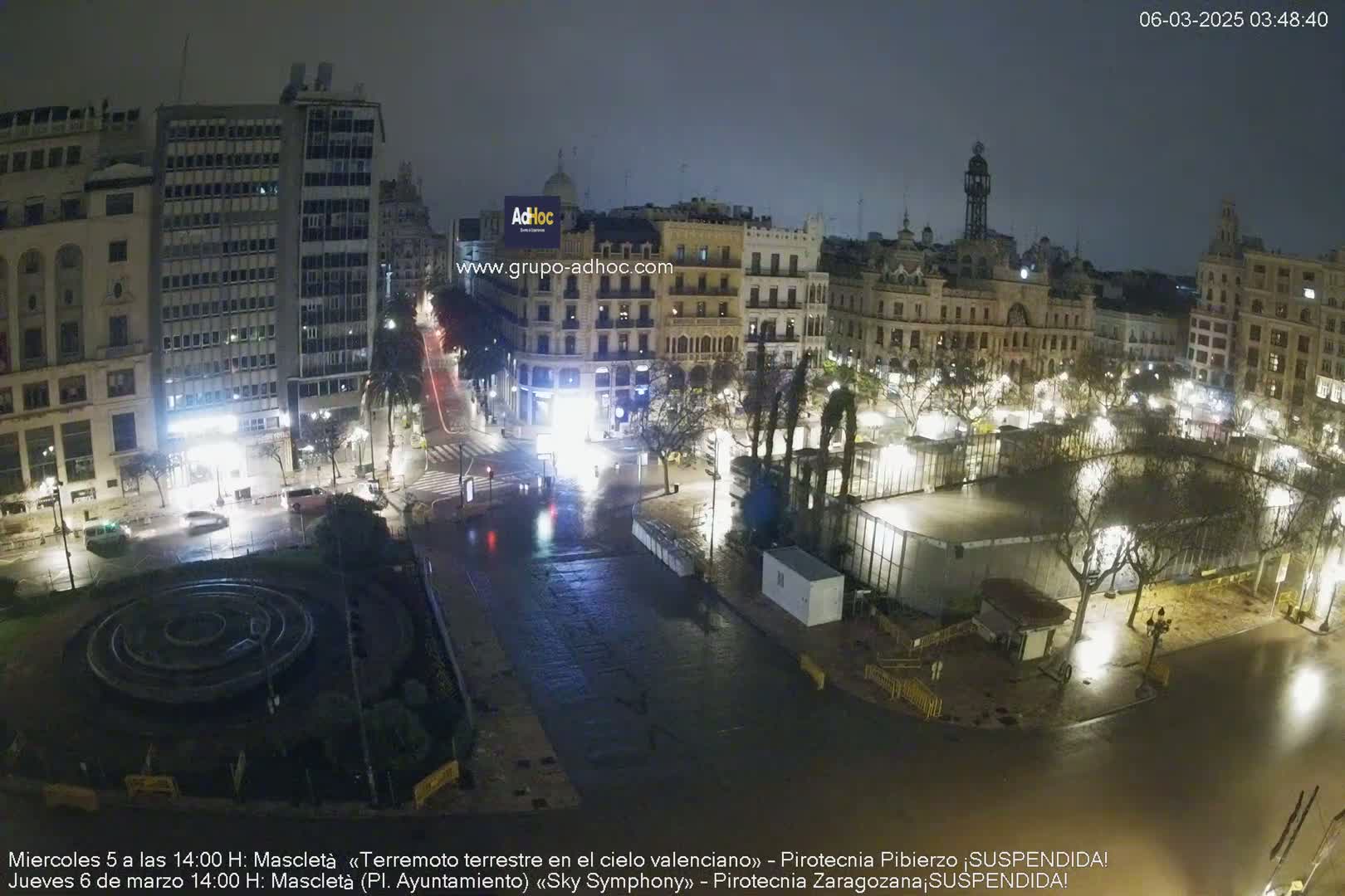A nighttime, wet city square scene shows several buildings and a circular fountain, with sparsely-lit streets and trees.