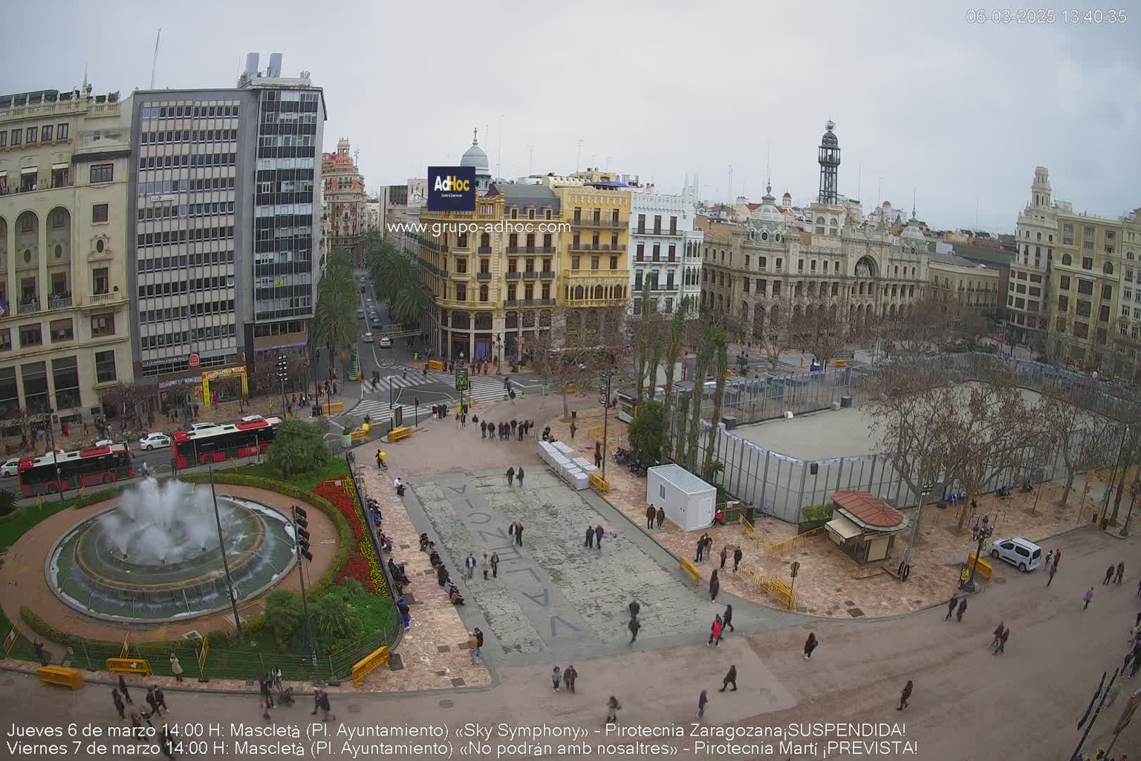 A city square with a fountain, numerous buildings, and sparse crowds under an overcast sky.