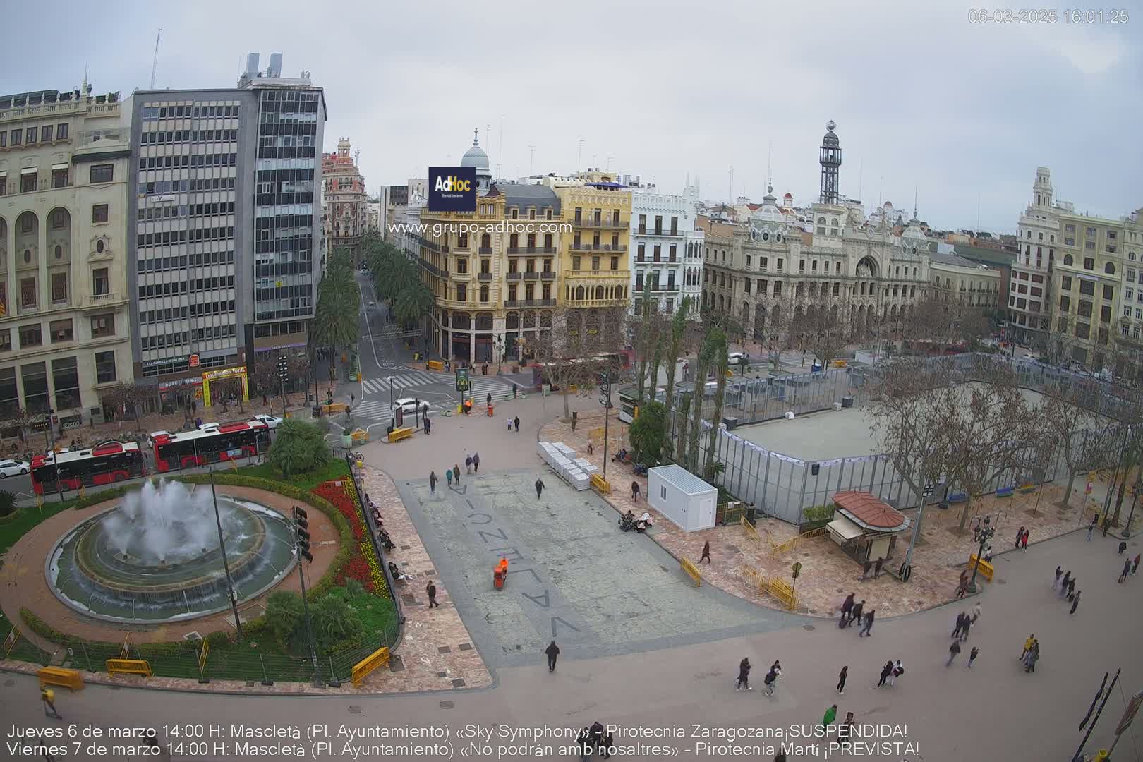 A city square with a fountain, buildings, and people under an overcast sky.