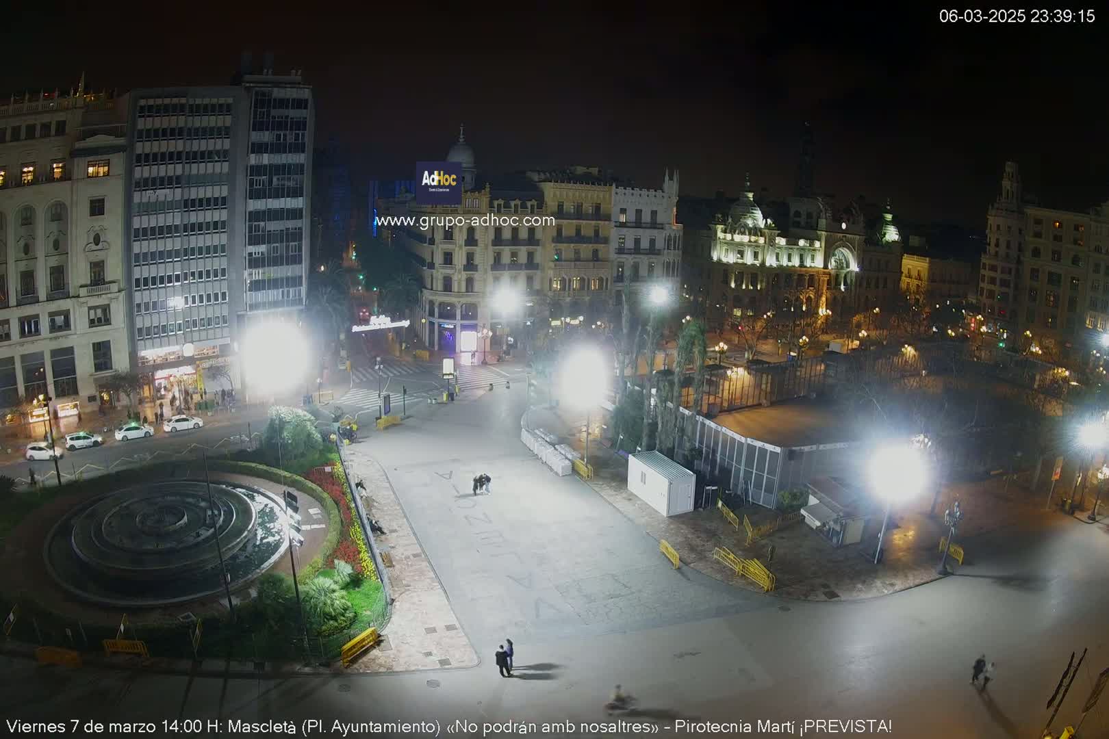 A nighttime, brightly lit city square with a fountain, buildings, and several people.