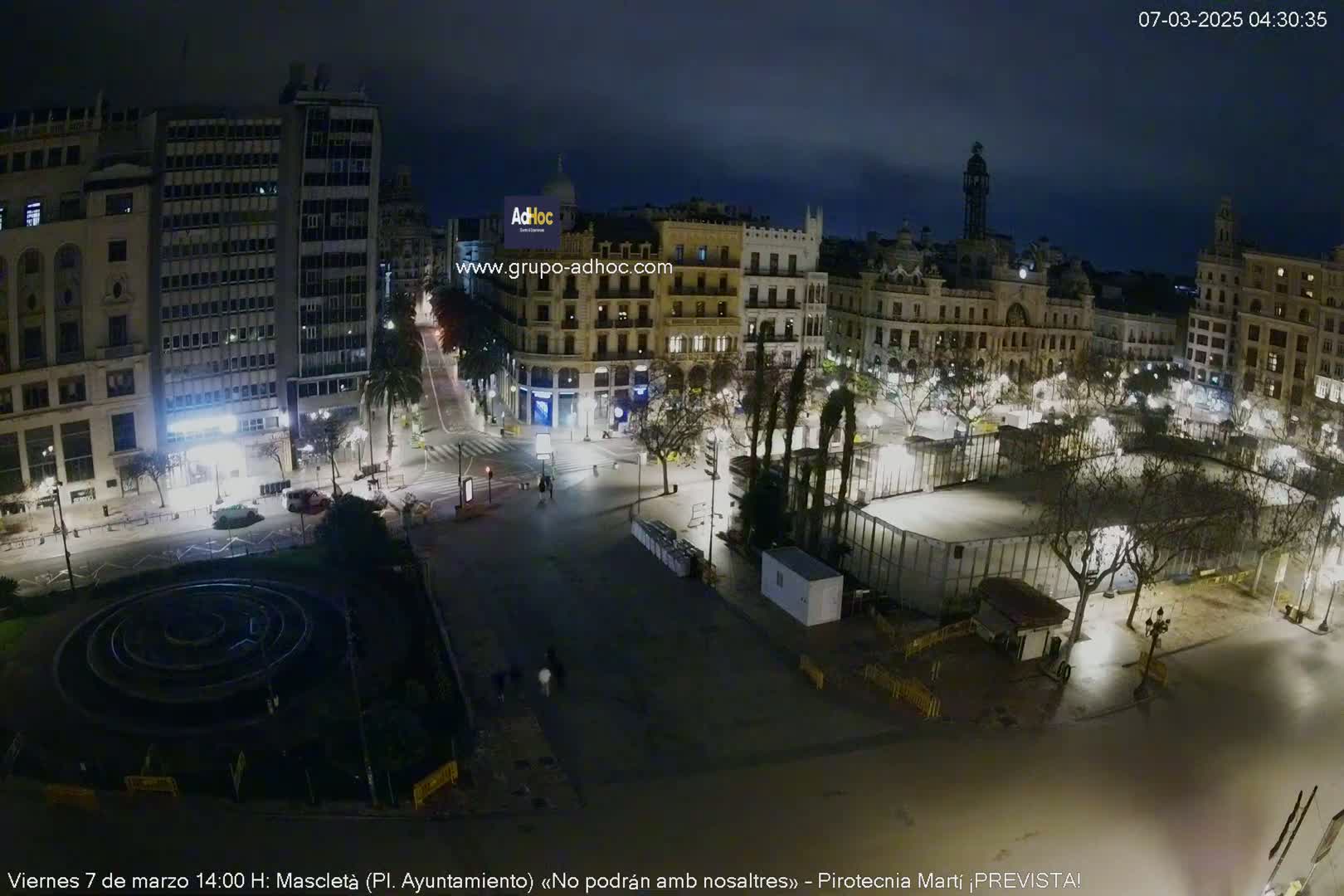 A nighttime, high-angle view of a city square with several buildings and trees, some of which are illuminated.