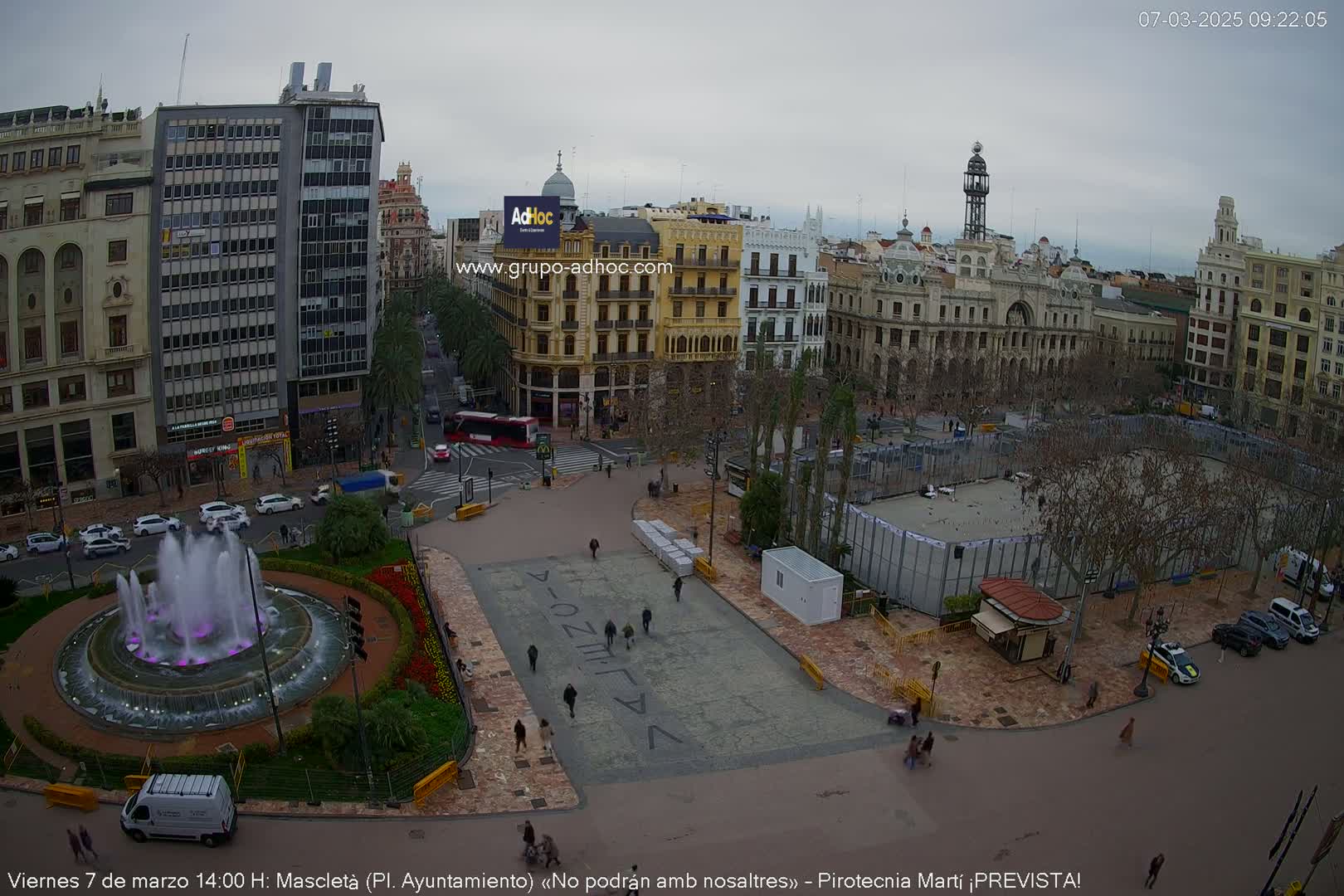 An aerial view of a city square on an overcast day shows a fountain, buildings, sparse pedestrian traffic, and a large fenced-off area in the center.