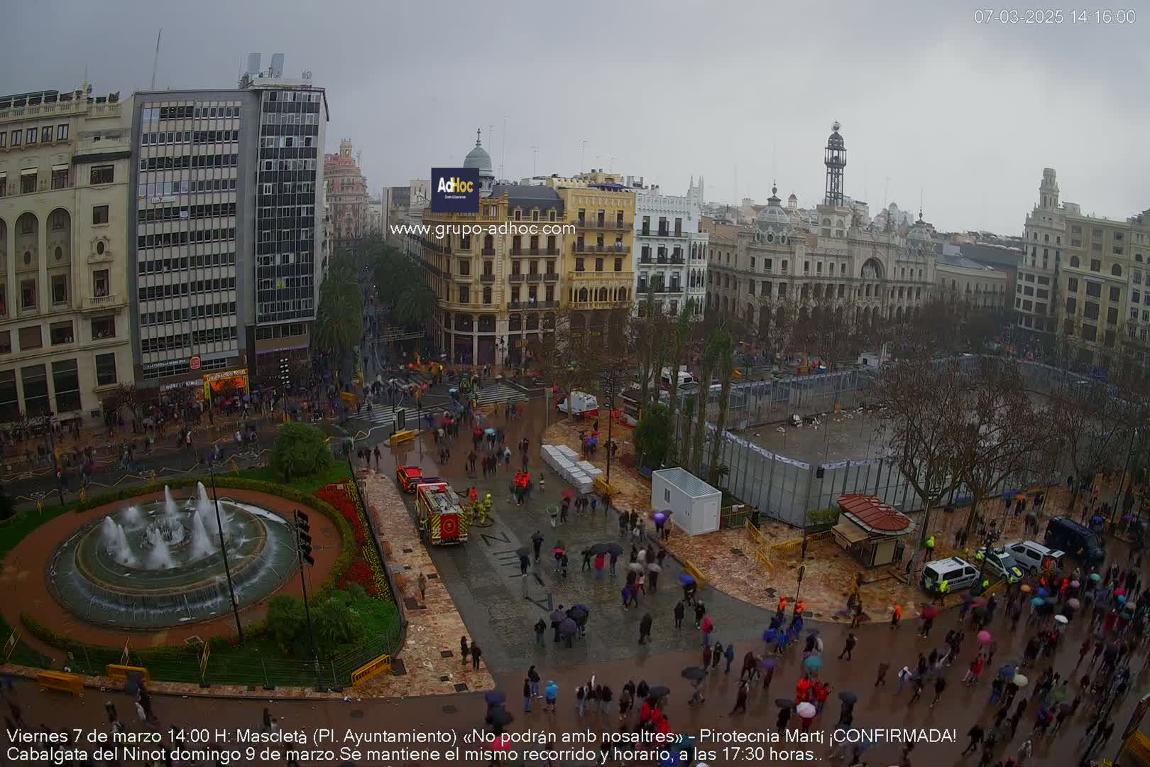 A rain-soaked city square, filled with many people under umbrellas, features a fountain, a firetruck, and temporary barriers surrounding a cleared central area.