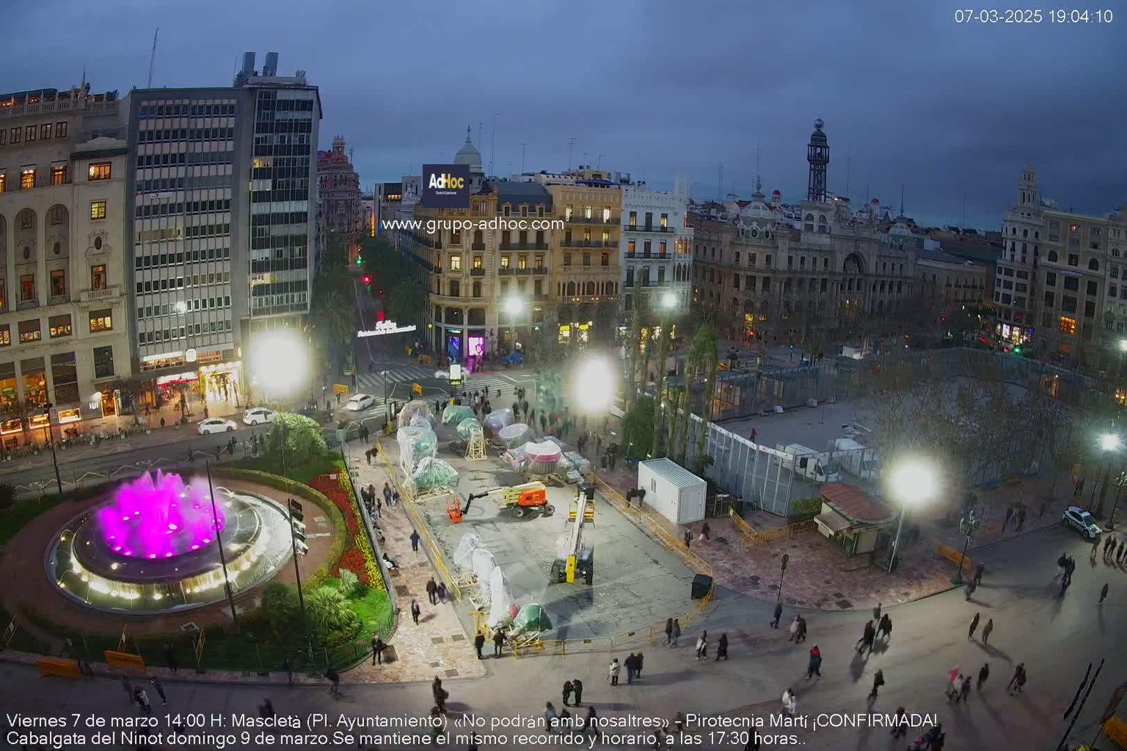 A city square at dusk is being prepared for an event, with construction equipment and barriers present around a central fountain lit with purple light.
