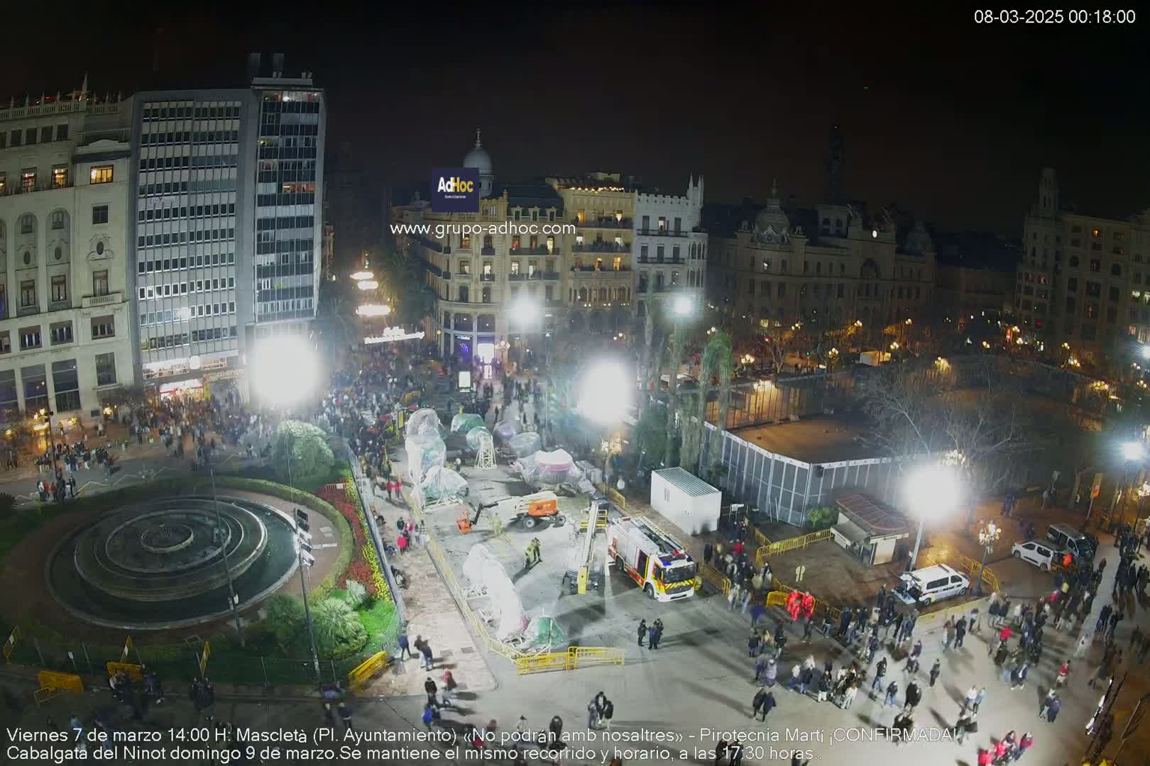 A nighttime aerial view of a city square with a crowd of people gathered around a central fountain and several vehicles, including a firetruck, under bright artificial lighting.