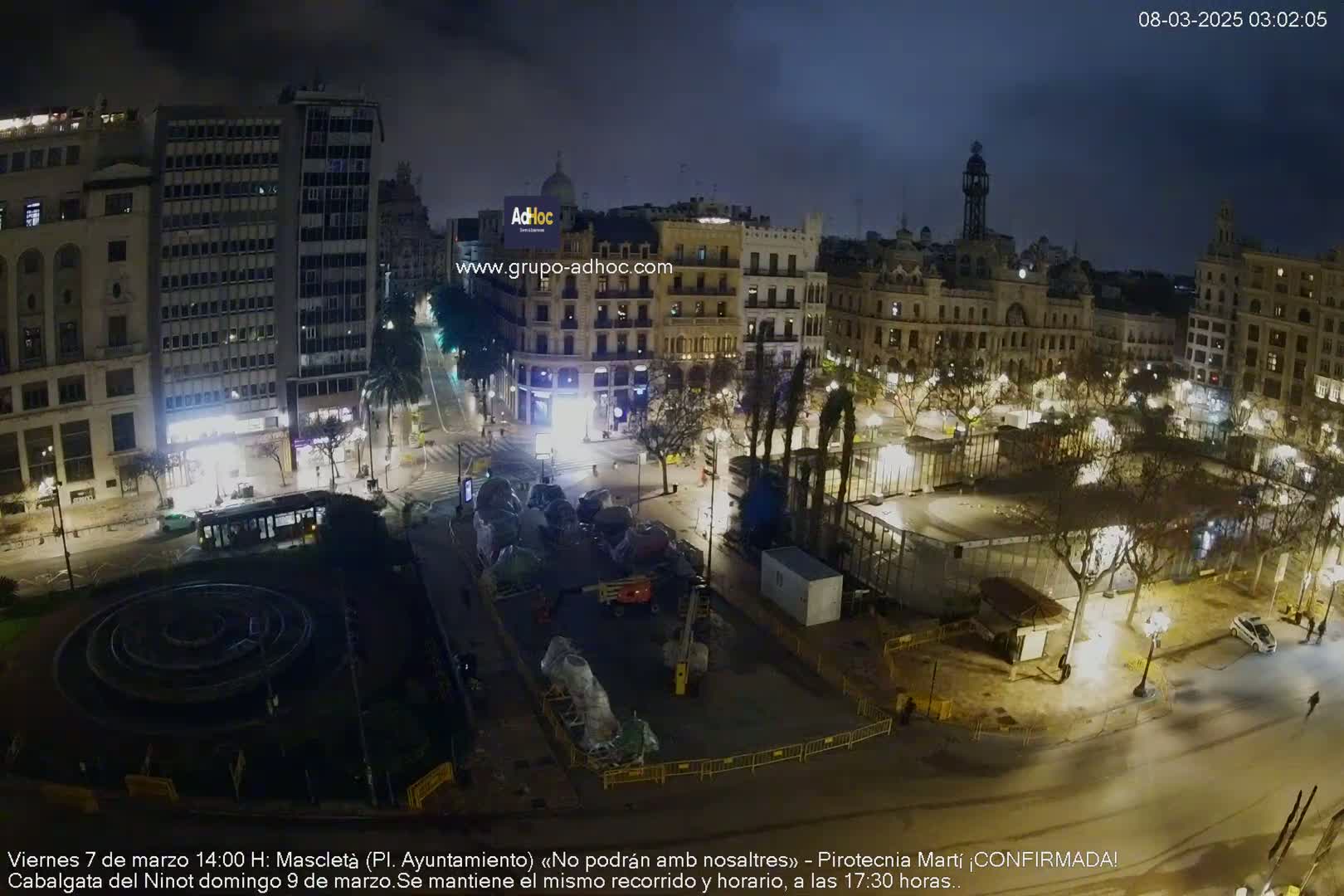 A nighttime aerial view of a city square with buildings lit up, a circular fountain, and construction or preparation activities underway.
