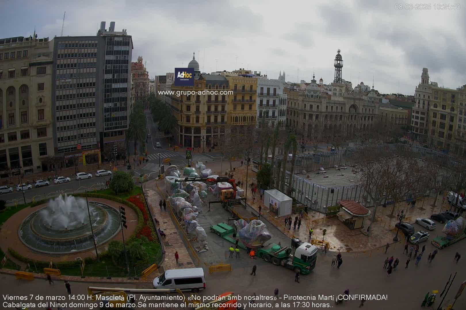 A city square is being prepped for an event, with numerous wrapped floats on flatbed trucks, a fountain in the foreground, and overcast skies.