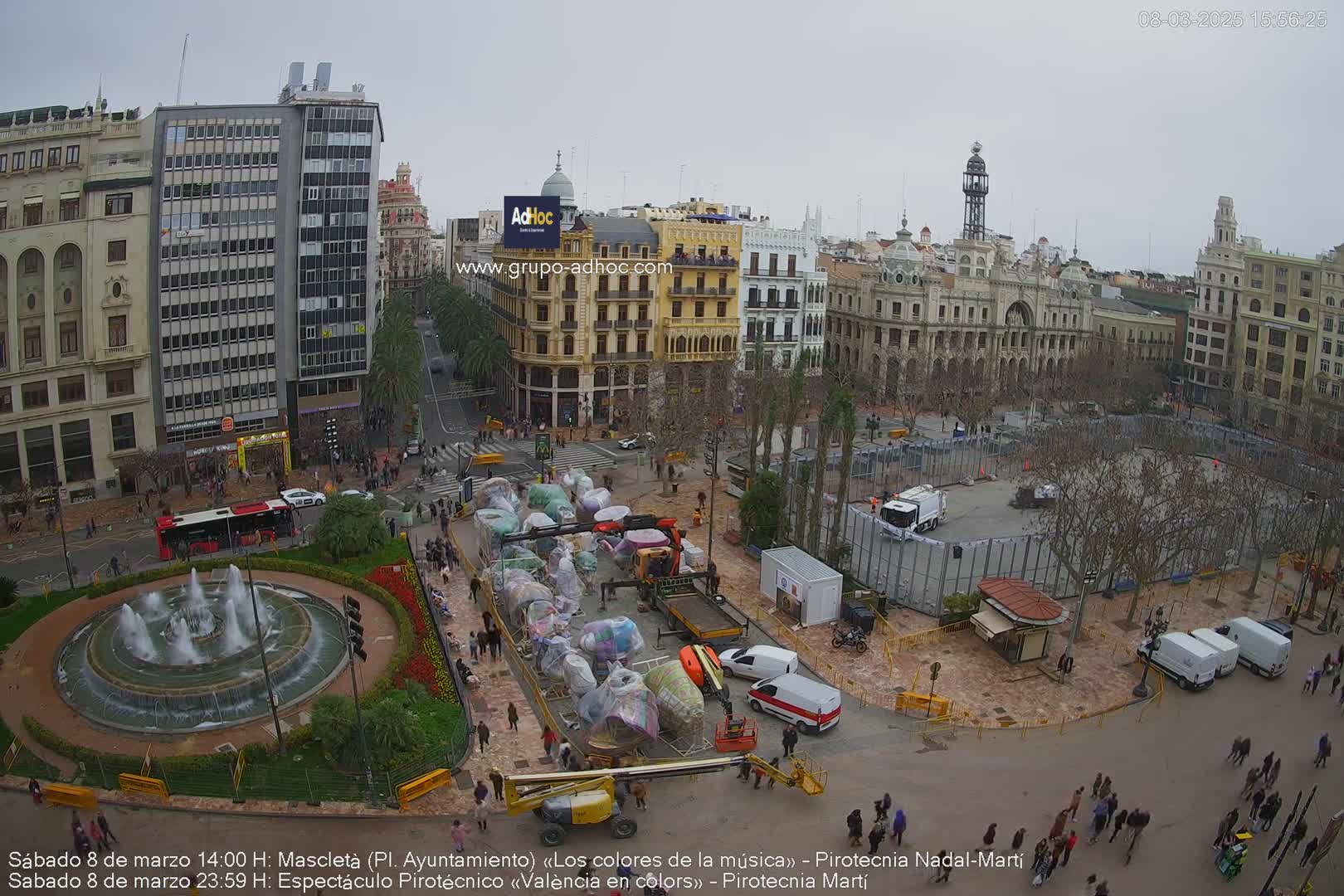 A city square is being set up for an event, with covered carnival rides and construction equipment present under an overcast sky.