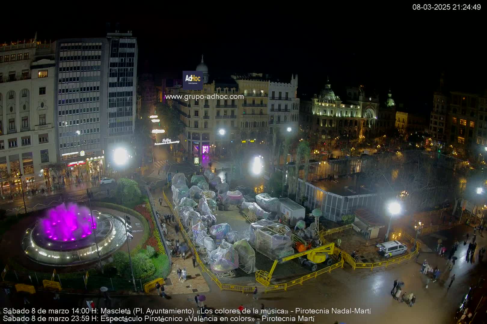 A nighttime aerial view of a city square shows a fountain with purple water, several covered carnival rides, and buildings under a clear sky.