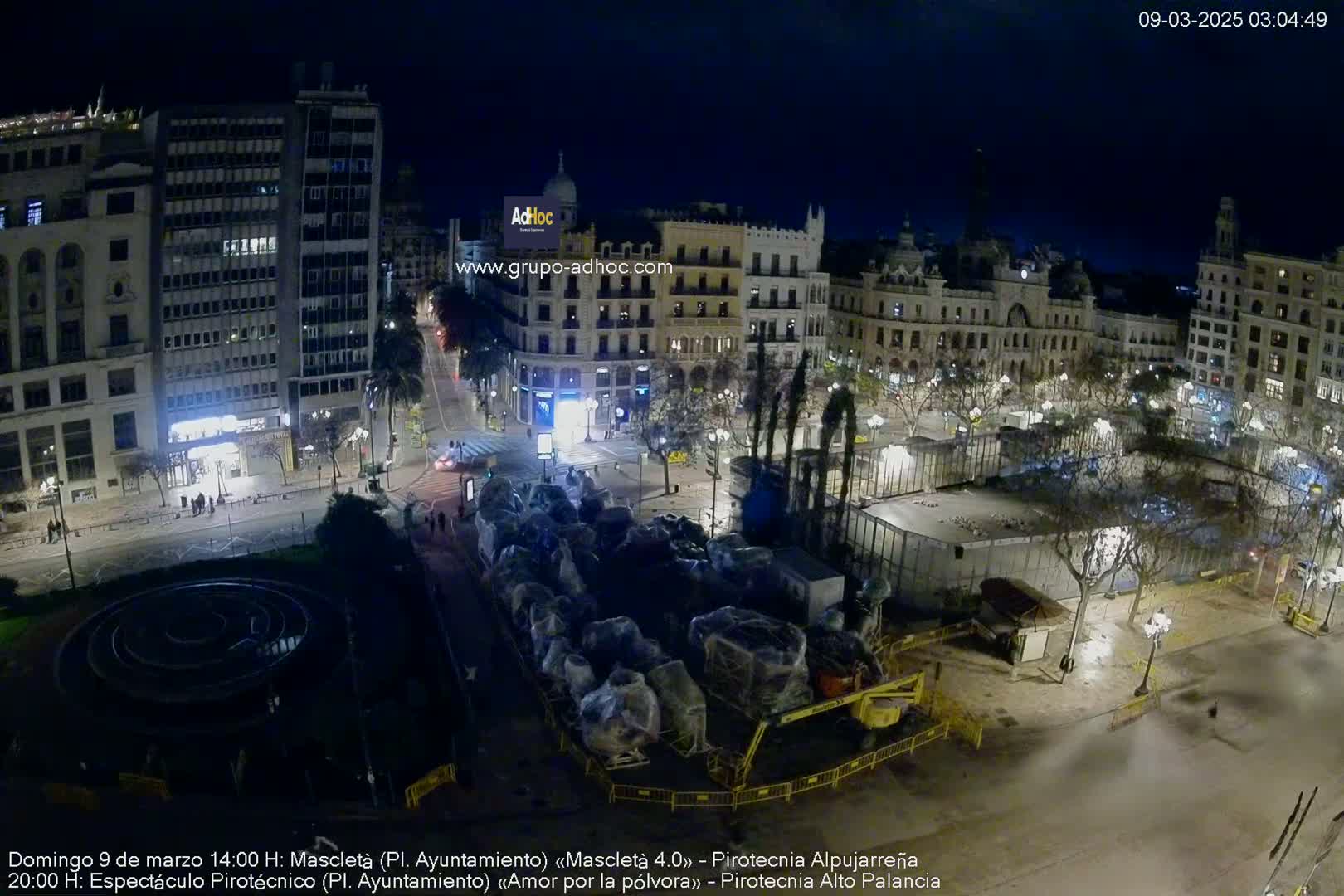 A nighttime aerial view of a city square with buildings surrounding it, showing preparations underway, possibly for a festival or event.
