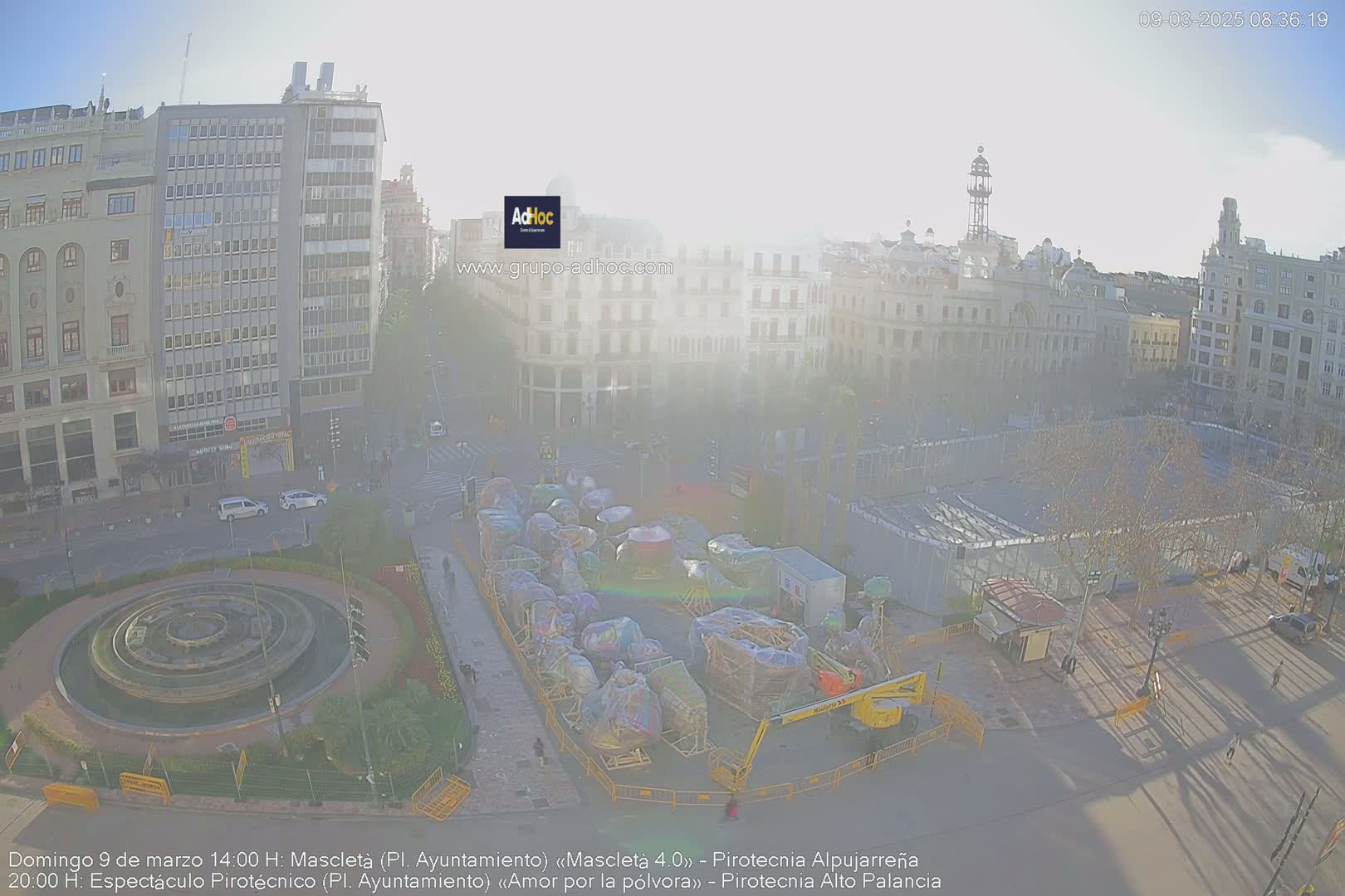 A hazy, sunny day reveals a city square with a circular fountain, numerous disassembled carnival rides, and buildings surrounding it.