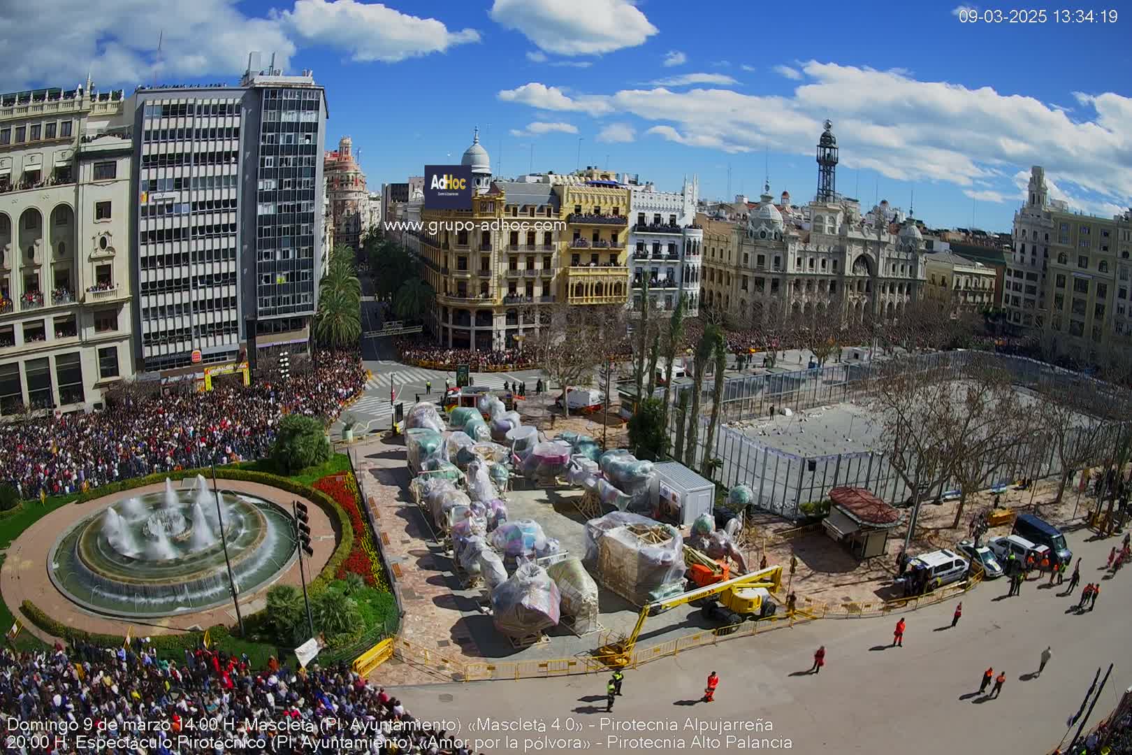 A large city square filled with a crowd of people, under a sunny blue sky with some clouds, features numerous covered floats and a central fountain.