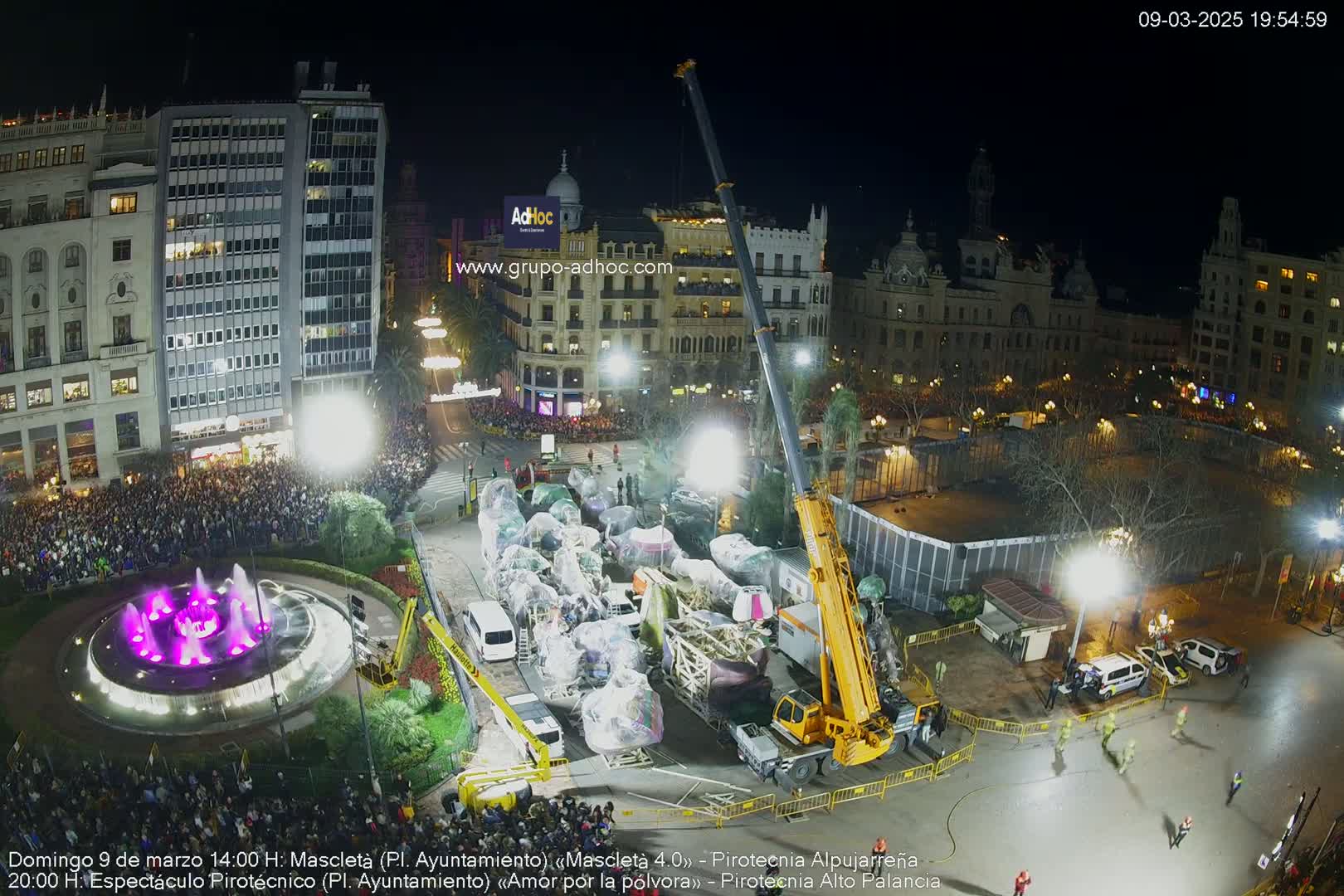 A large crowd gathers in a city square at night around a fountain illuminated in purple, with a yellow crane lifting what appear to be covered floats near several vehicles.