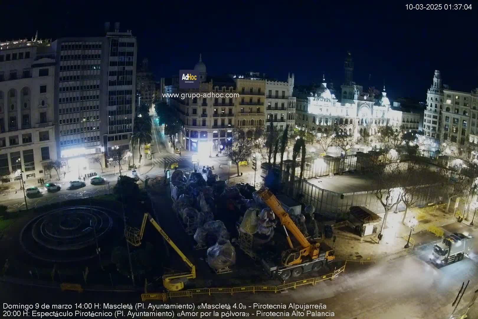 A nighttime aerial view of a city square with buildings illuminated, a crane lifting large objects in the center, and a circular fountain visible in the foreground.