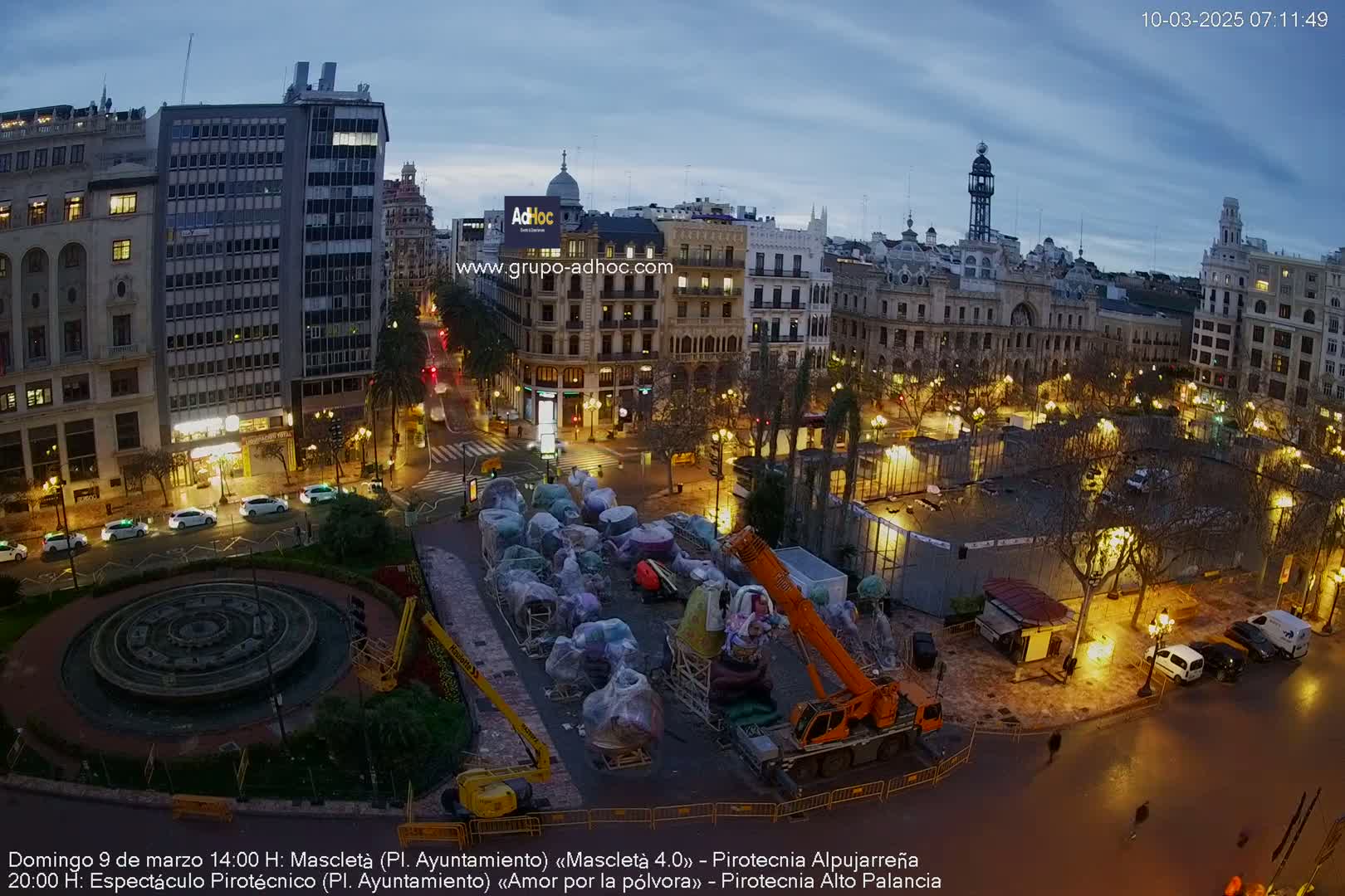 A city square at dusk under a cloudy sky, showing a crane lifting what appears to be large, wrapped sculptures in front of illuminated buildings and a fountain.
