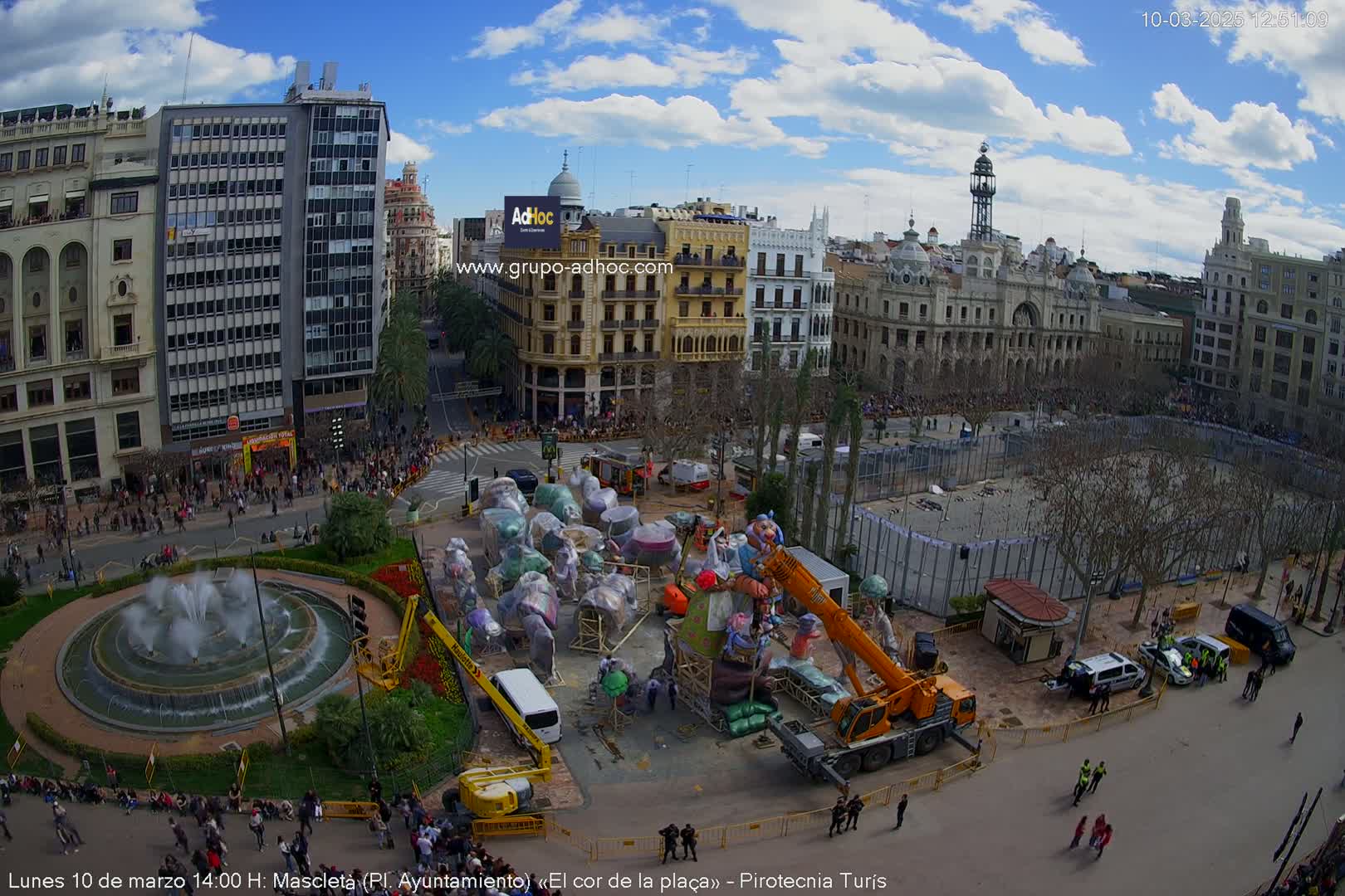 A city square under a partly cloudy sky, featuring a large fountain, numerous people, and a crane lifting large, covered structures near several buildings.