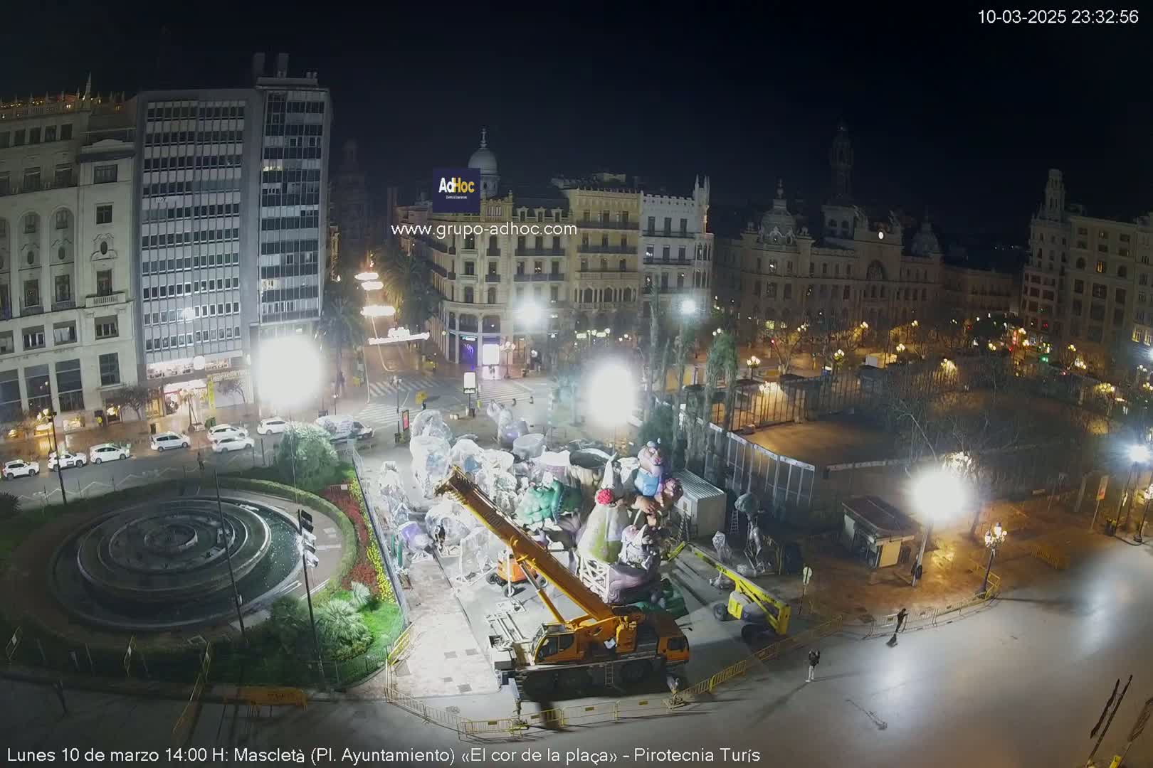 A nighttime, brightly lit city square features a crane lifting large, covered figures near a circular fountain, with buildings and trees surrounding the square.