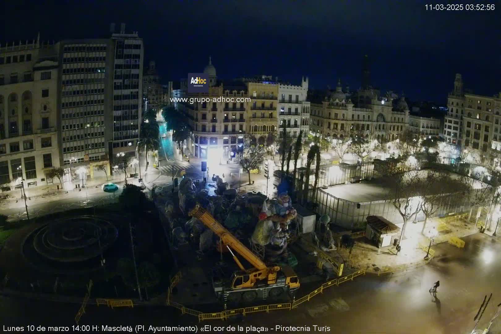 A nighttime, aerial view of a city square with a crane lifting a large sculpture, surrounded by buildings and trees illuminated by streetlights.