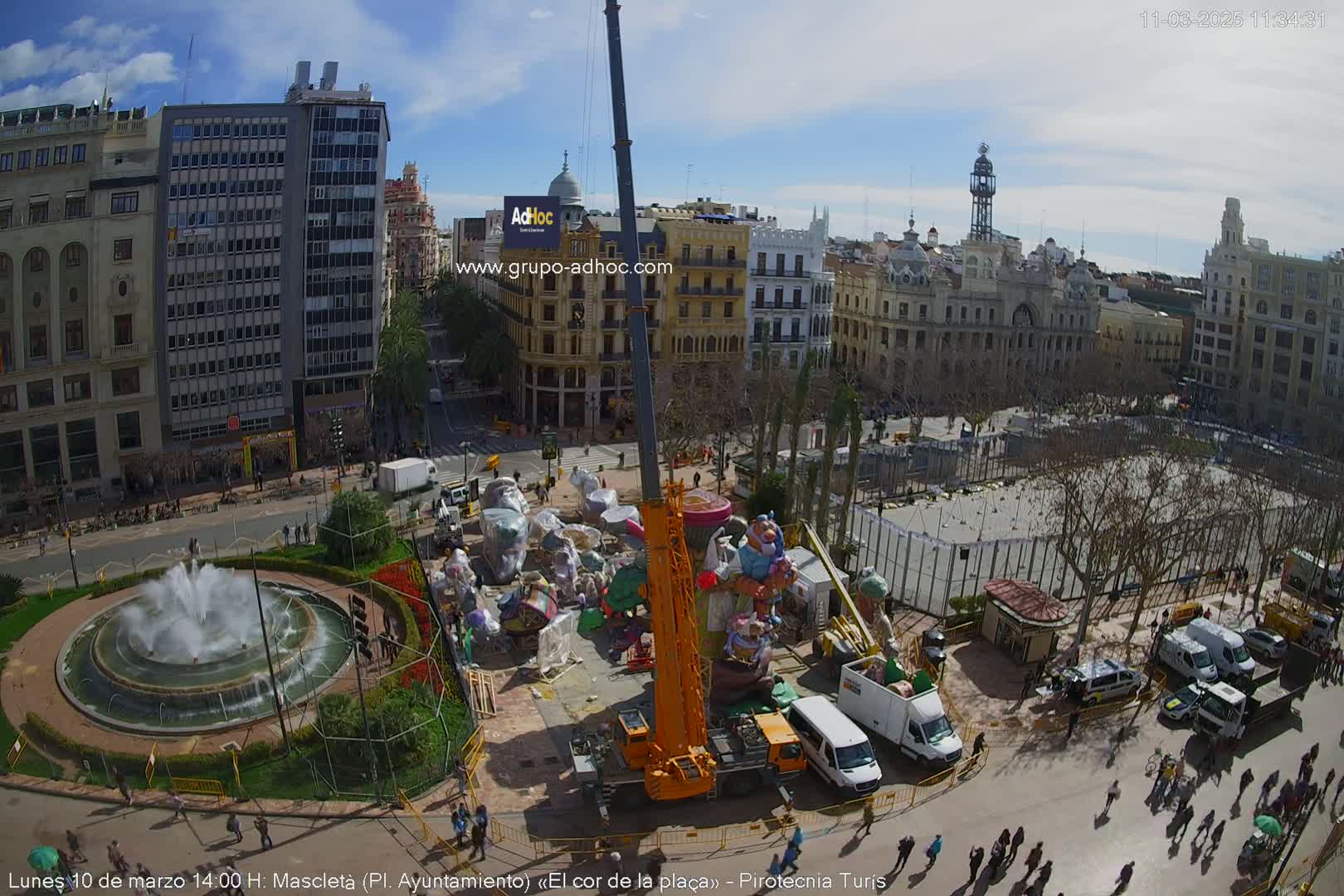 A crane lifts a large, colorful structure in a city square under a mostly sunny sky, with people walking around the perimeter and smaller structures nearby.