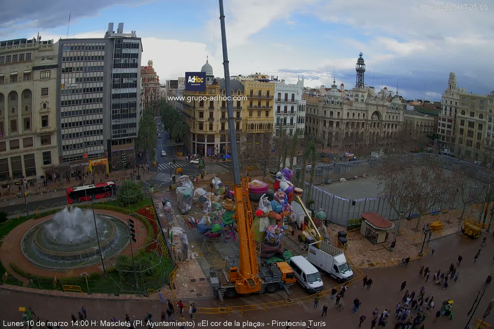 A city square is shown under a partly cloudy sky, with a large crane lifting a colorful display in the center, surrounded by people and buildings.