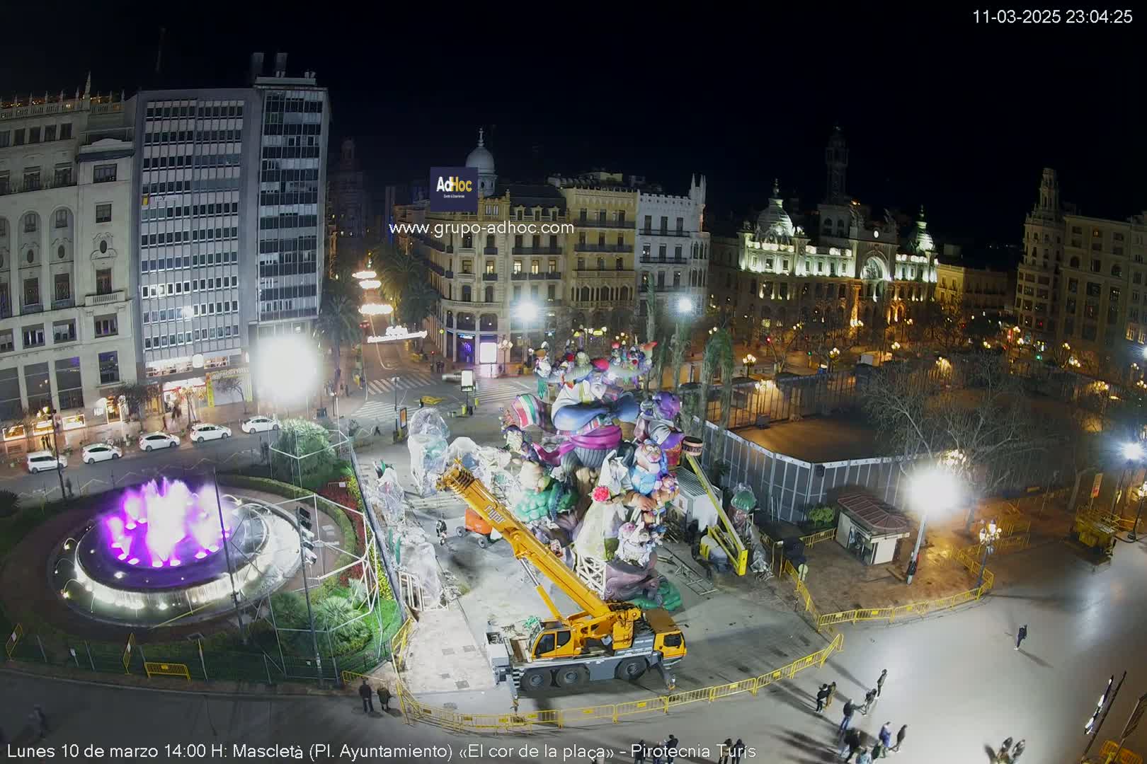 At night, a large crane lifts a colorful, whimsical sculpture in a city square with a lit fountain and surrounding buildings.