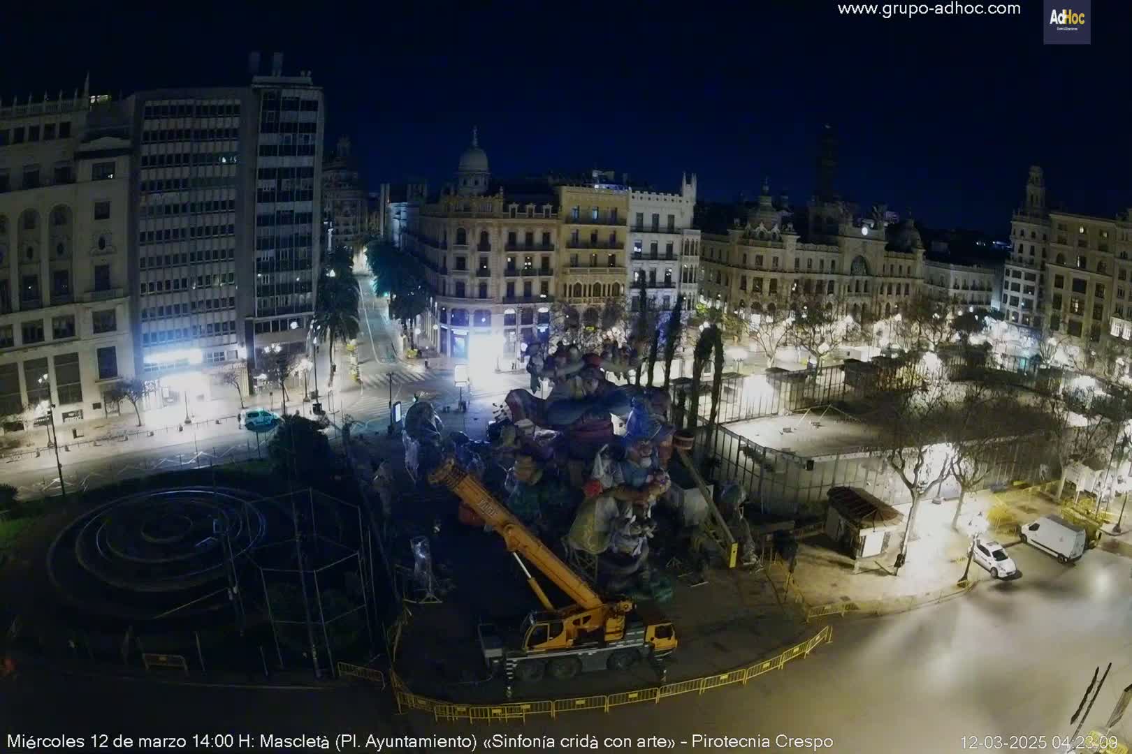A nighttime aerial view of a city square shows a large sculpture being lifted by a crane, surrounded by buildings and trees under a clear, dark sky.