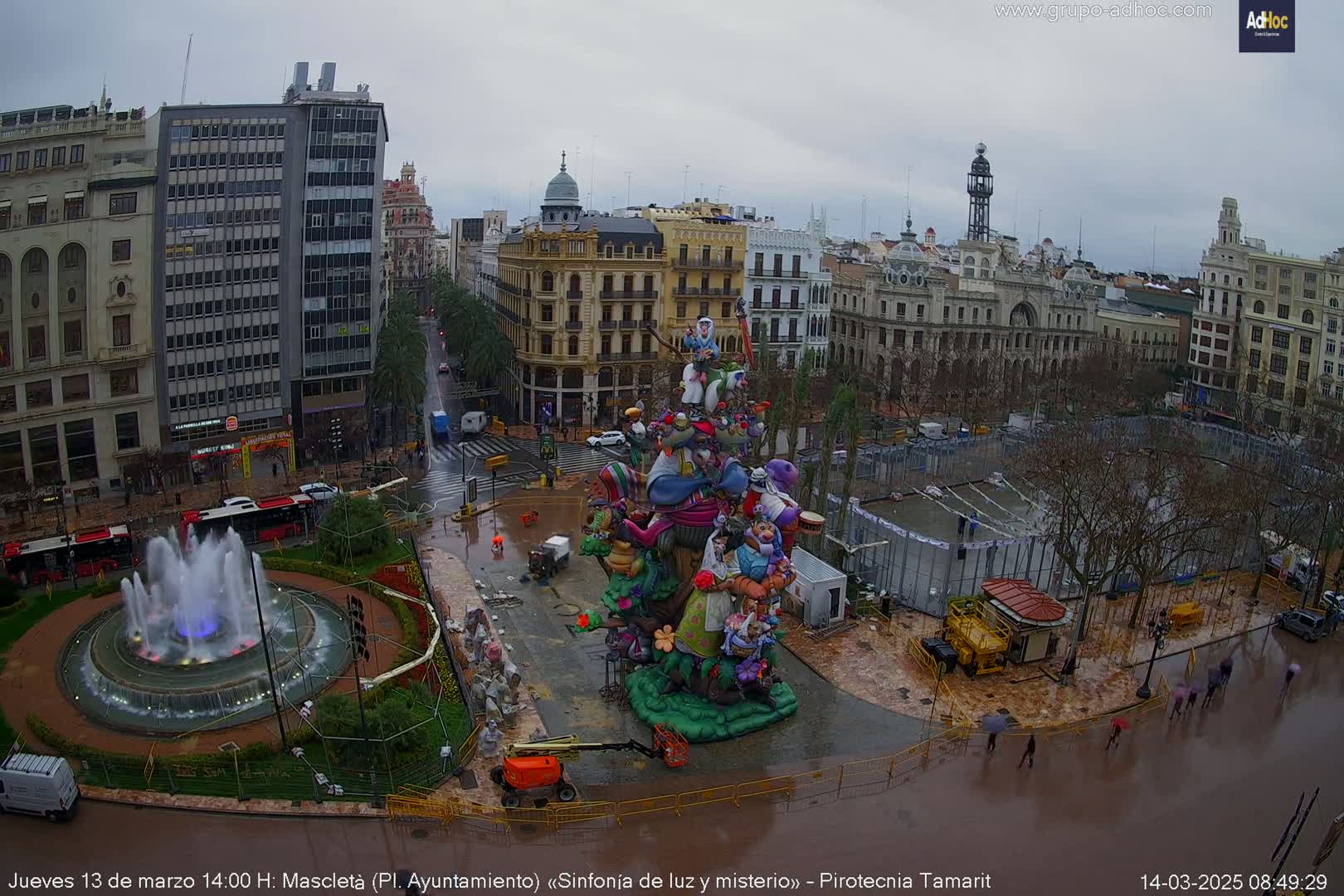A large, colorful sculpture sits in a city square during a rainy day, surrounded by buildings, a fountain, and people walking under umbrellas.