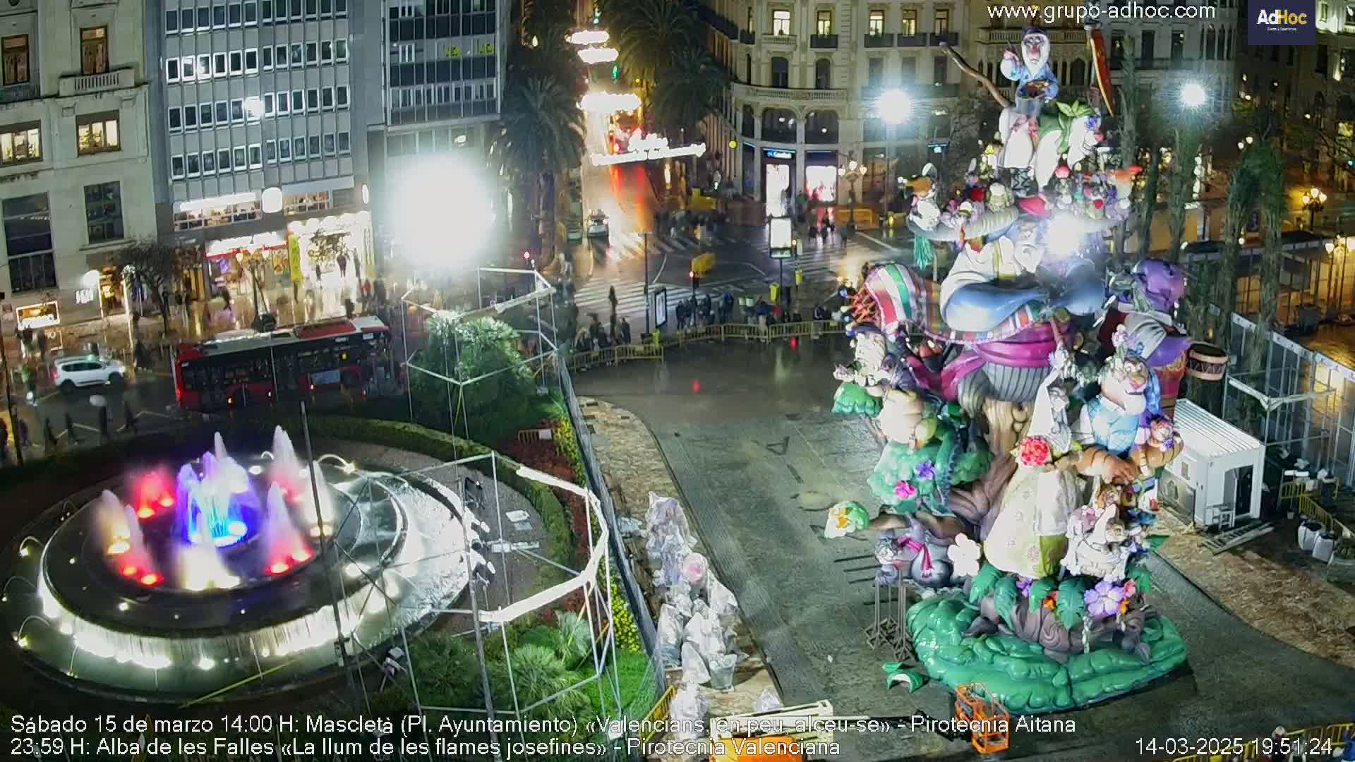 A nighttime view of a city square featuring a large, elaborate float or sculpture, a circular fountain with colorful lights, and a wet, illuminated street scene.