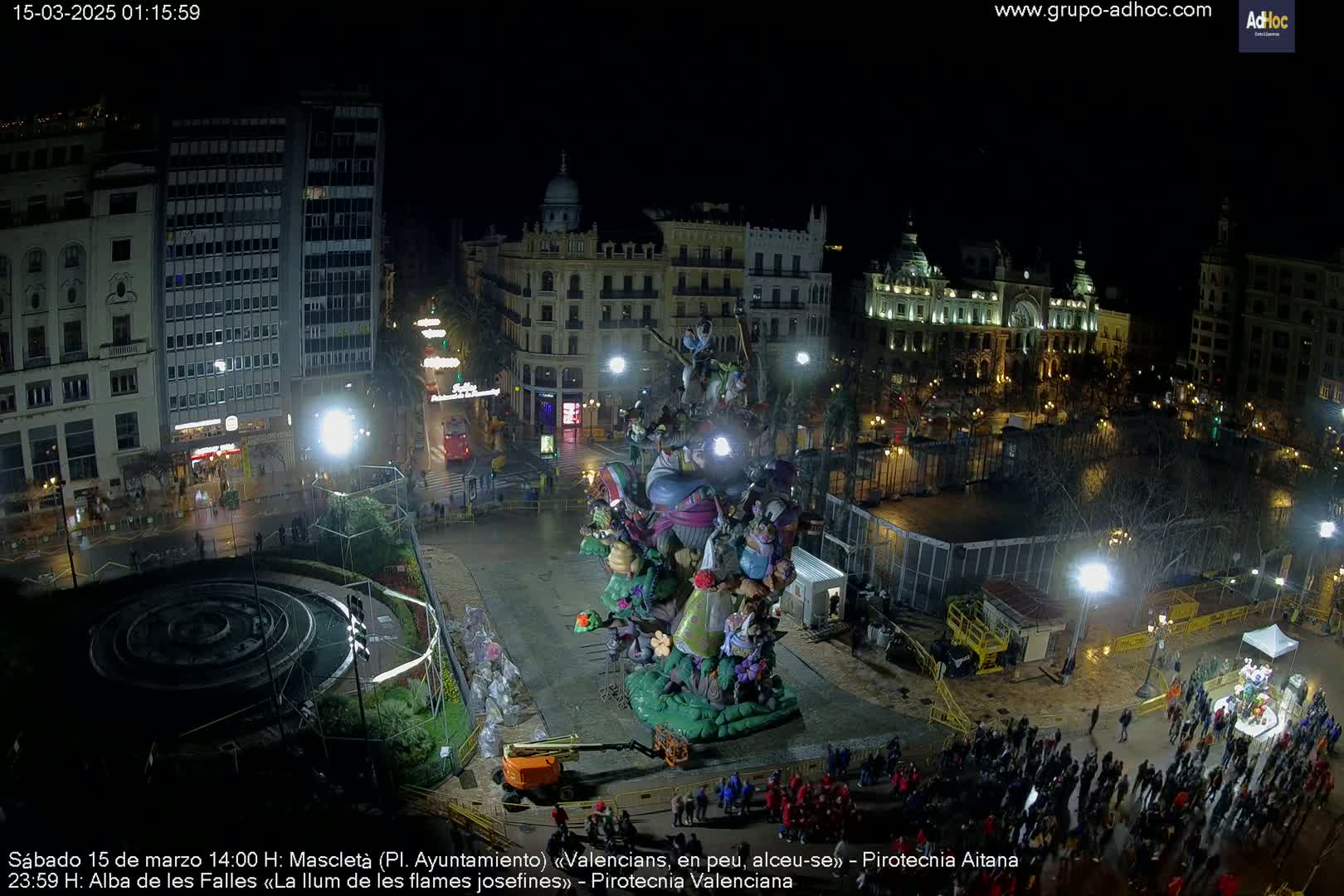 A nighttime aerial view of a city square with a large, elaborate, colorful statue at its center, surrounded by people and illuminated by artificial lights.
