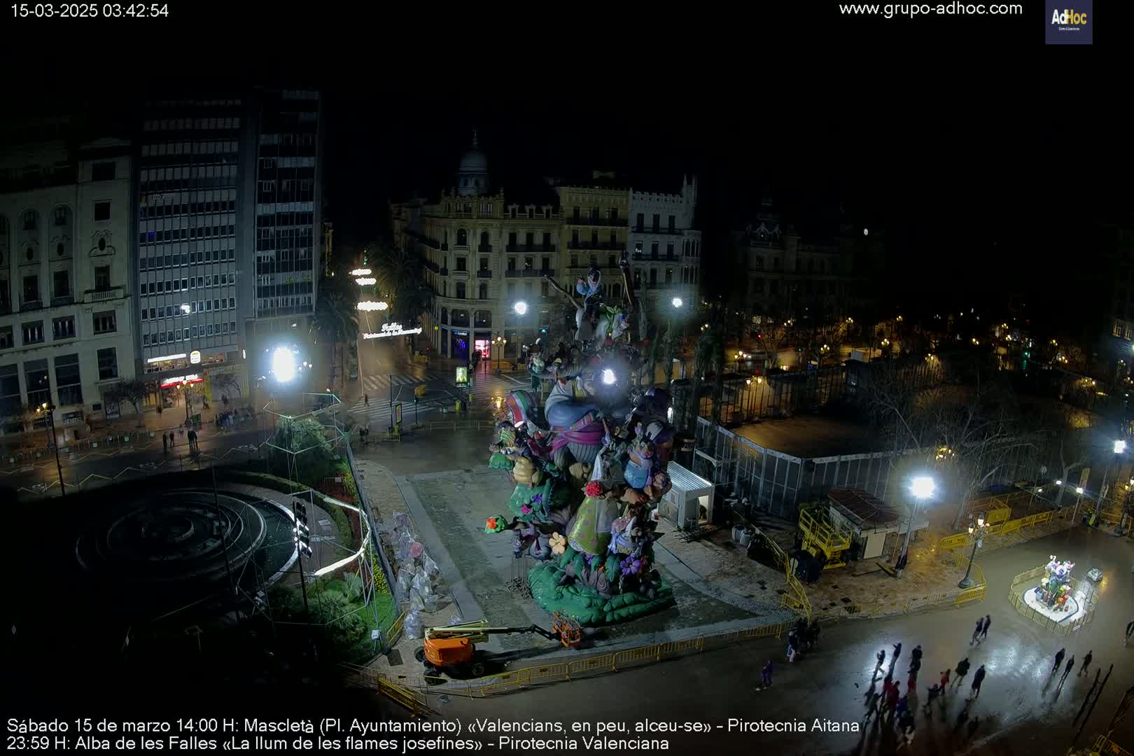 A nighttime, aerial view of a large, colorful statue in a city square, surrounded by buildings and people.