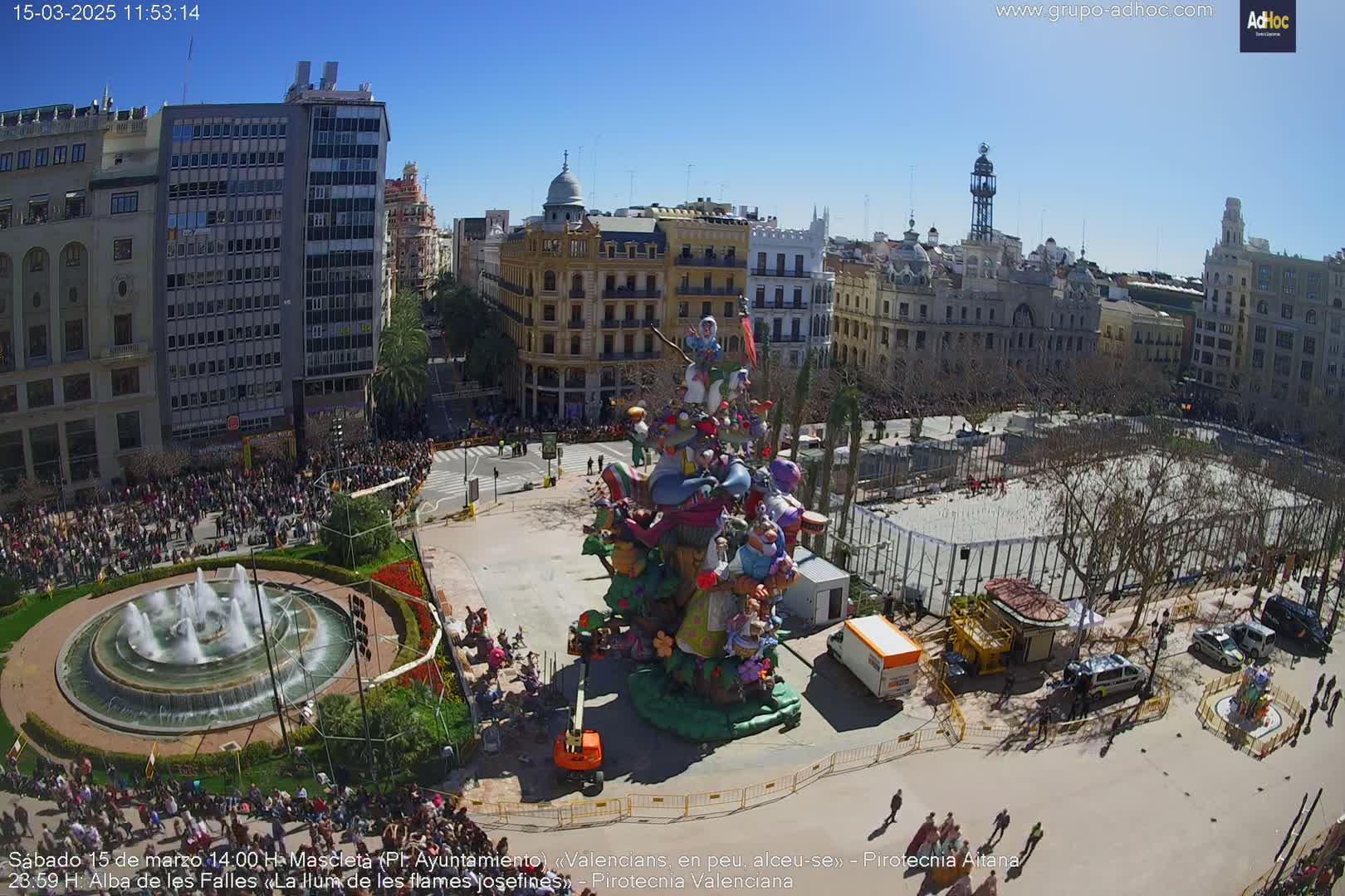 A large crowd surrounds a colorful, elaborate float in a city square on a sunny day.