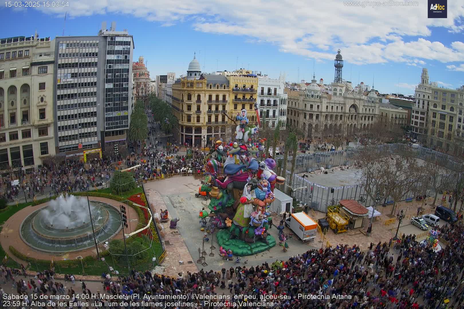 A large crowd surrounds a colorful, elaborate sculpture in a city square under a partly cloudy sky.