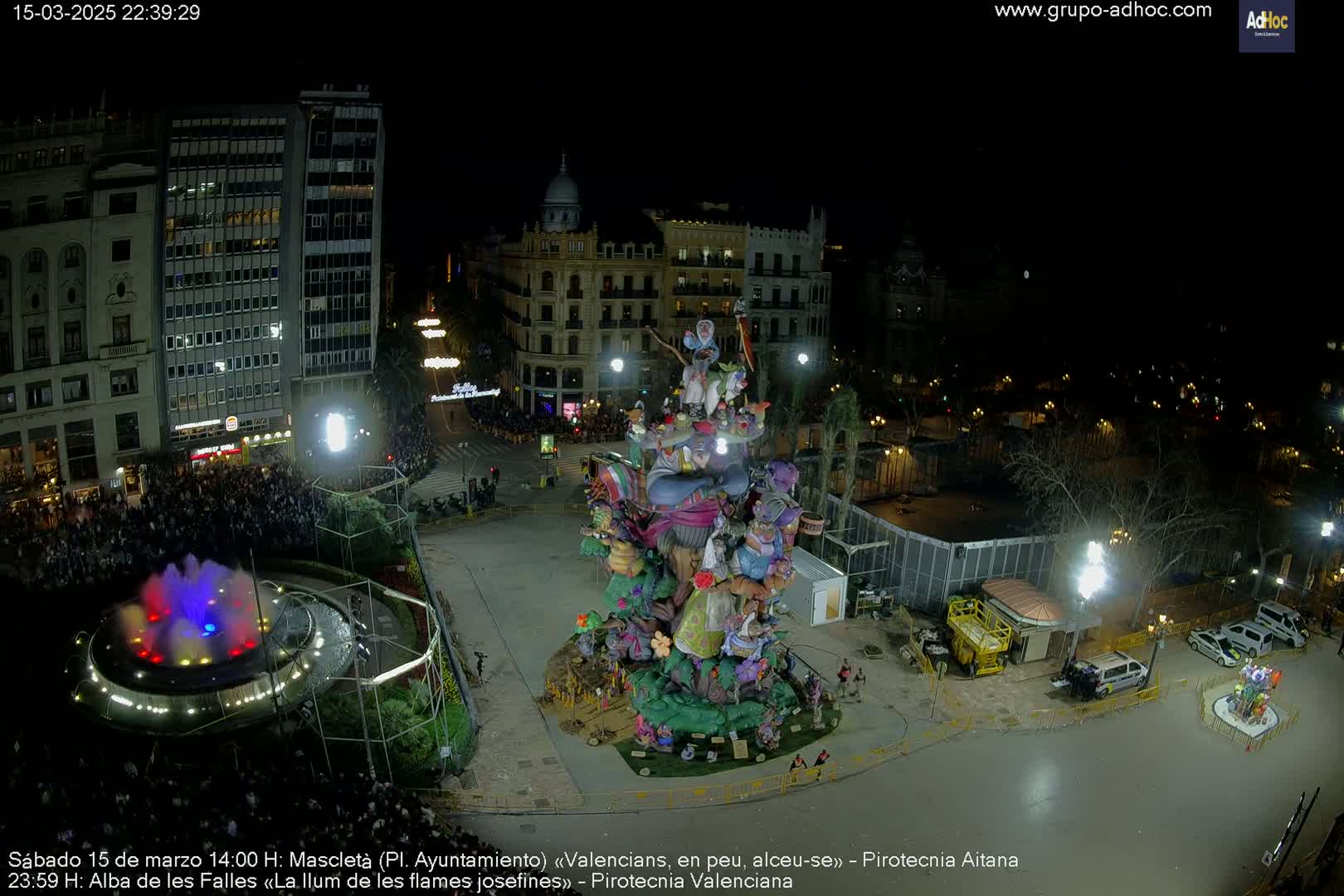 A nighttime aerial view shows a large, colorful, whimsical sculpture in a city square surrounded by buildings and a crowd of people, with a fountain in the foreground.