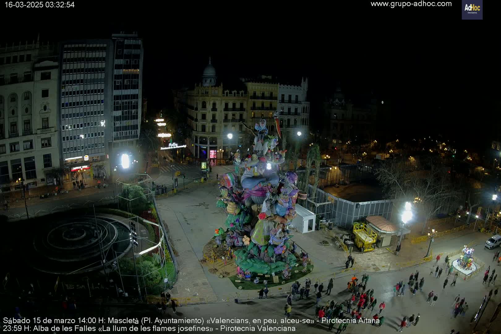 A large, colorful, nighttime display featuring numerous figurines stands in a city square, surrounded by people and buildings under dark, clear skies.