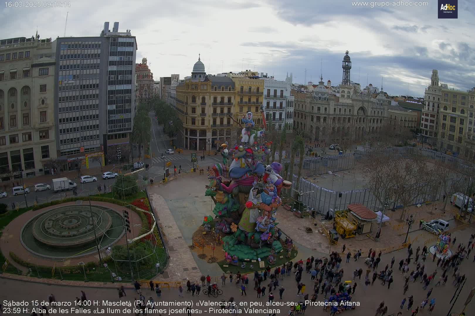 A large, colorful, whimsical sculpture stands in a city square under a cloudy sky, surrounded by people and buildings.