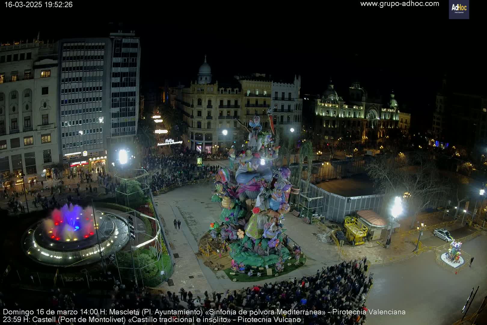 A nighttime aerial view of a city square with a large, colorful, ornate structure in the center, surrounded by a crowd of people, a fountain, and buildings illuminated by lights.