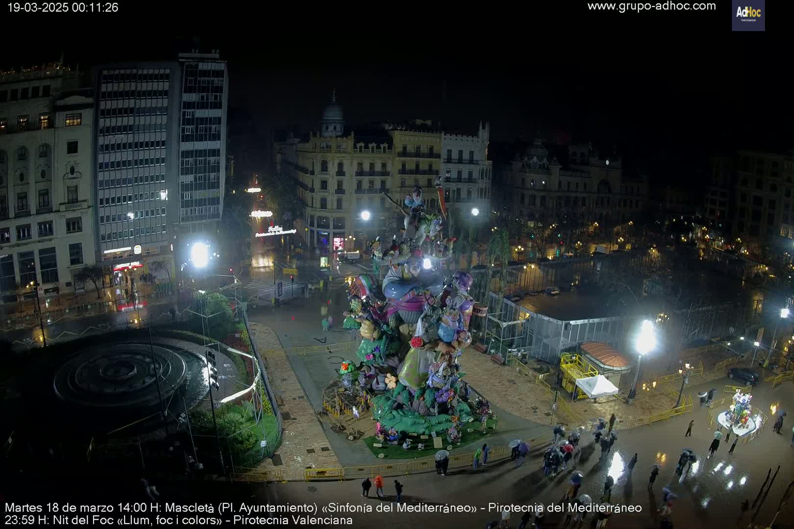 A nighttime, rain-soaked city square features a large, elaborate, colorful sculpture in the center, surrounded by people under umbrellas.