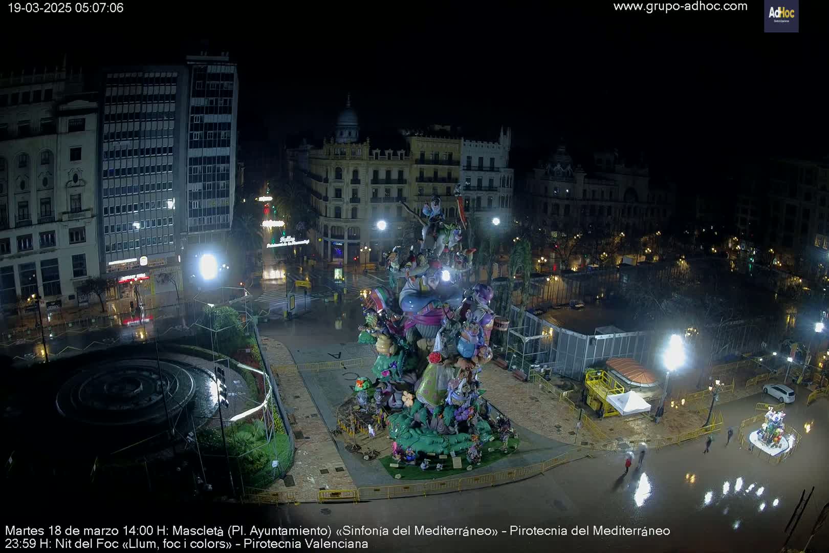 A large, colorful, intricately designed sculpture stands in a rain-soaked plaza at night, surrounded by city buildings and illuminated by streetlights.