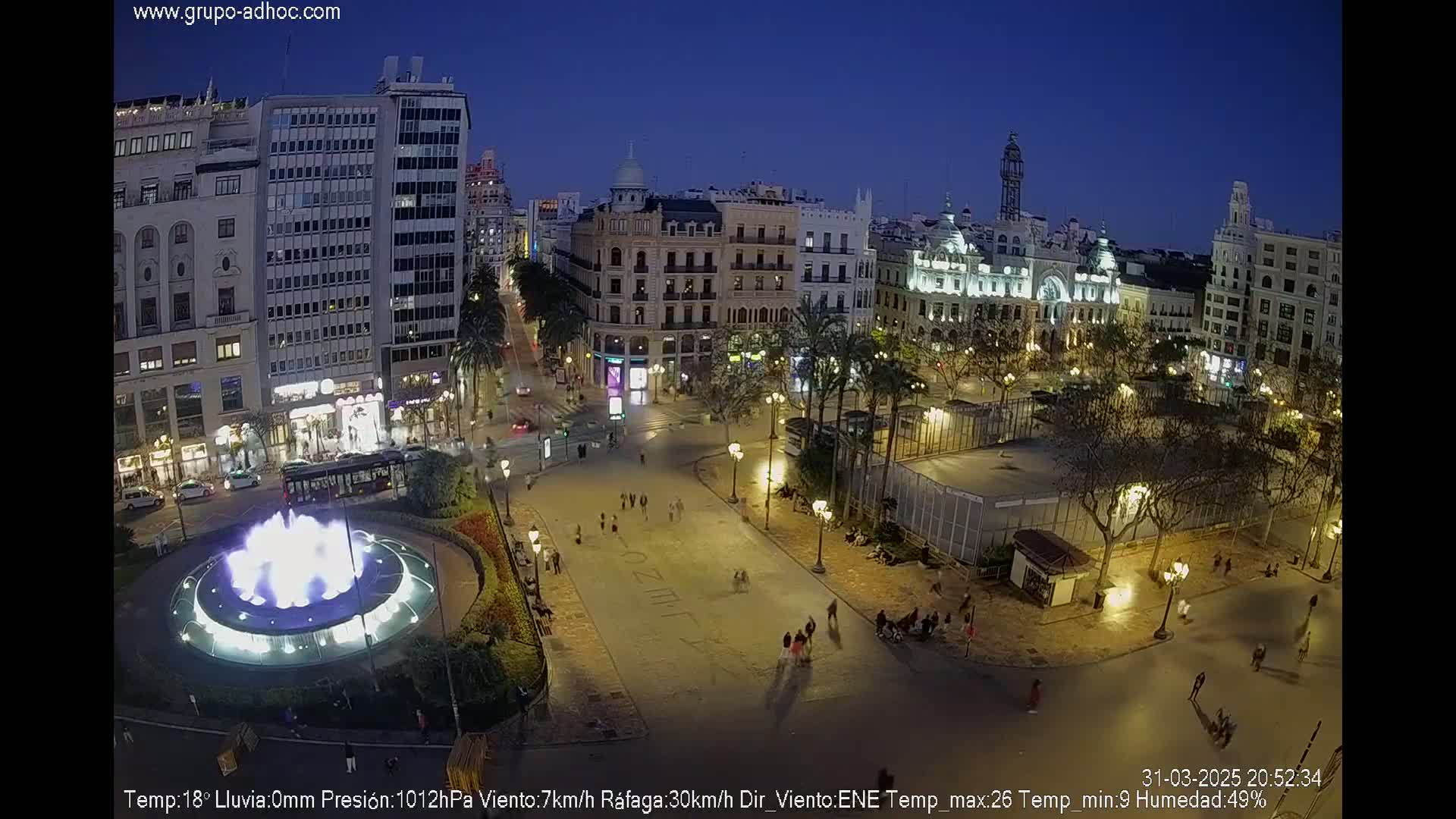 A city square at night, illuminated by streetlights, features a central fountain and is surrounded by buildings under a clear, dark blue sky.