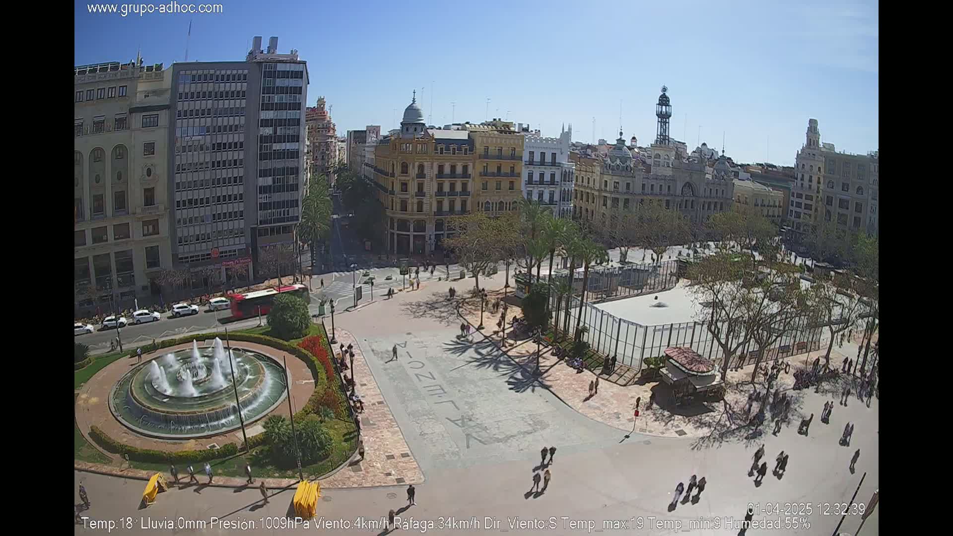 A high-angle view of a city square on a sunny day, featuring a circular fountain, palm trees casting shadows, and buildings surrounding the open space.