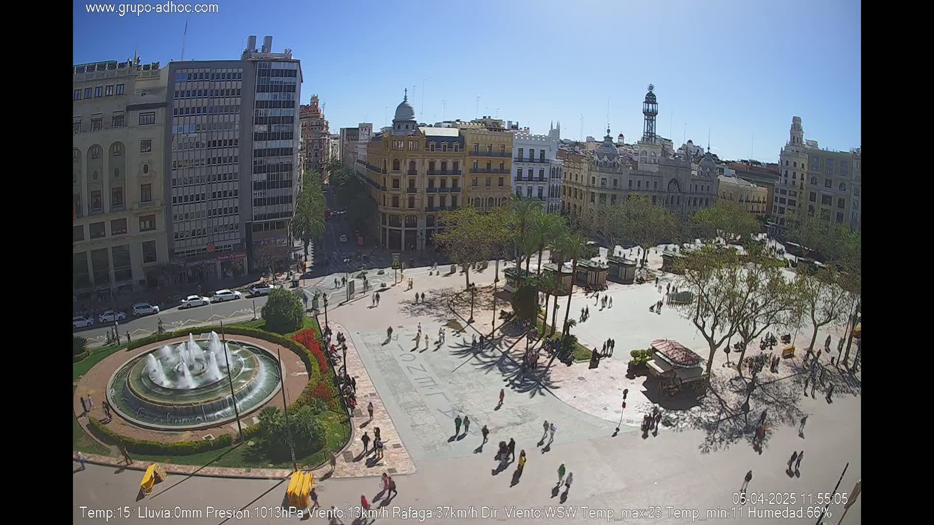 A sunny city square with a central fountain, surrounded by buildings and palm trees, with people walking around.