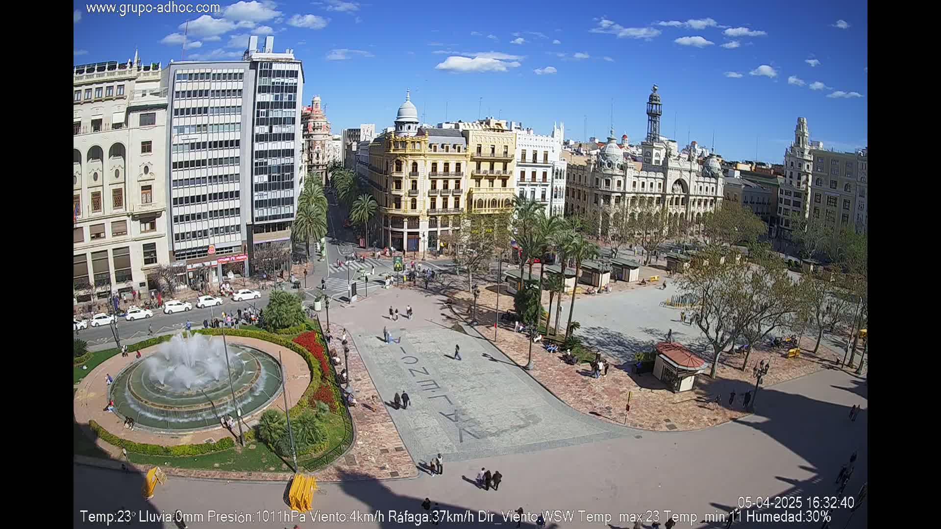 A sunny city square with a fountain, surrounded by buildings and palm trees, under a mostly clear blue sky.