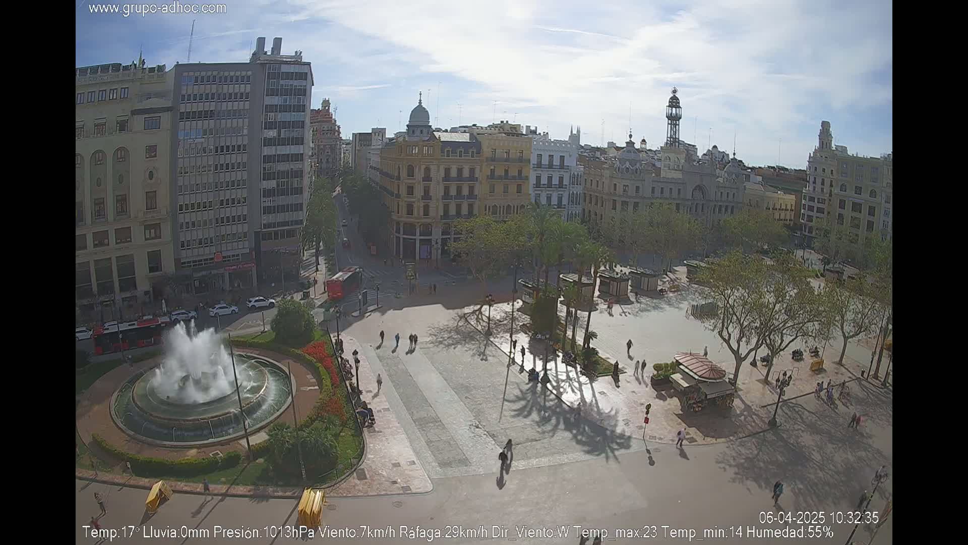 A sunny city square with a central fountain, surrounded by buildings and trees, with people walking around.