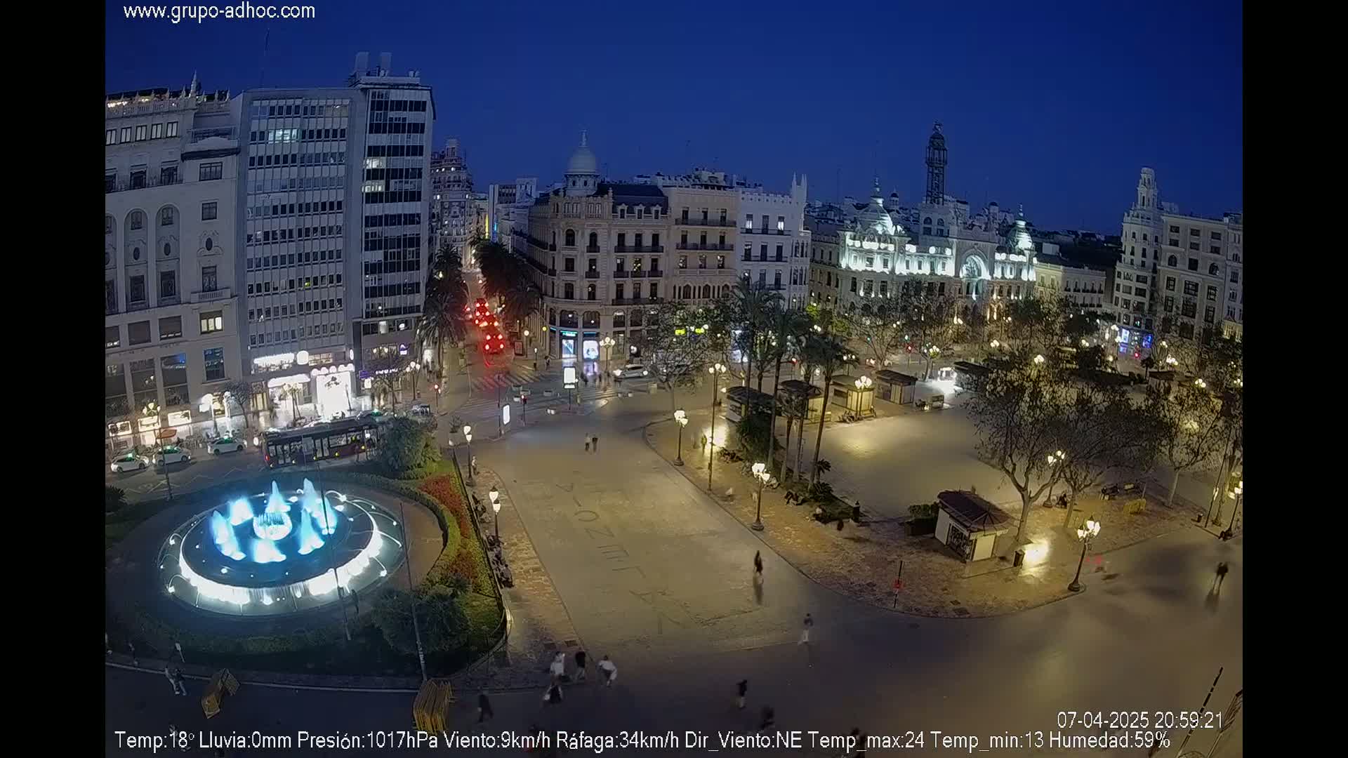 A nighttime view of a city square with a fountain, illuminated buildings, and sparsely populated streets under a dark blue, clear sky.