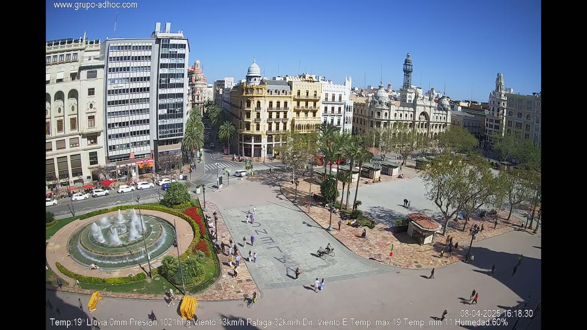 A city square with a fountain, surrounded by buildings and palm trees, on a sunny day.