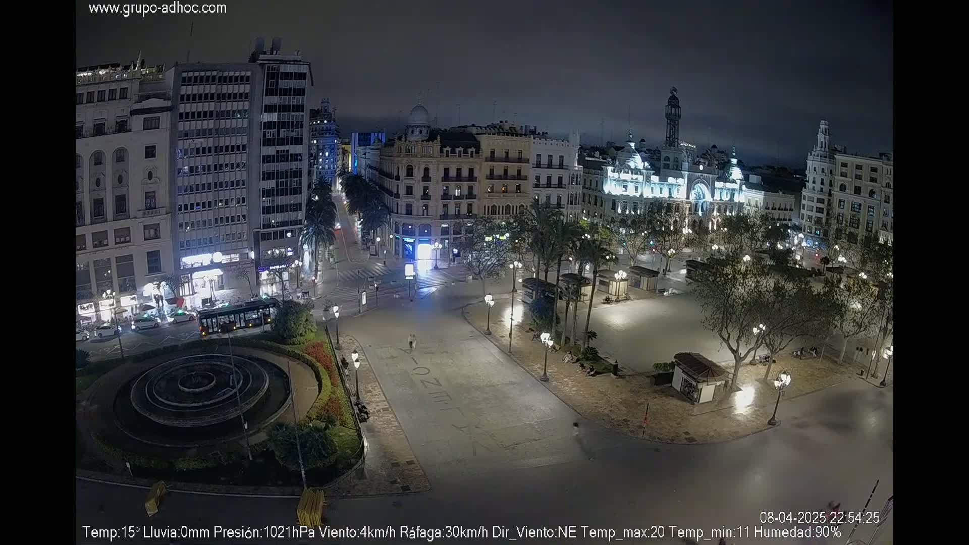 A nighttime, mostly empty city square with a fountain and surrounding buildings, under a clear sky.
